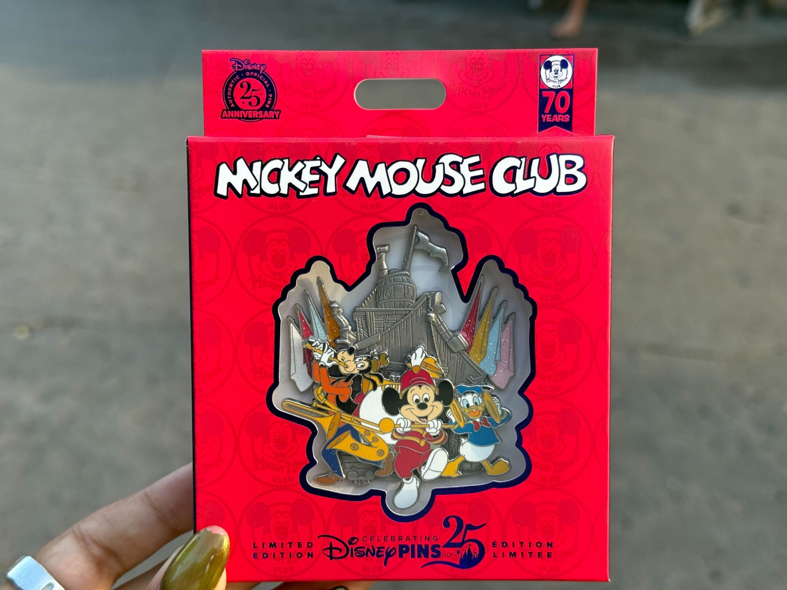 A hand holds a Disney “Mickey Mouse Club” 25th anniversary limited edition pin in front of a blurred background.