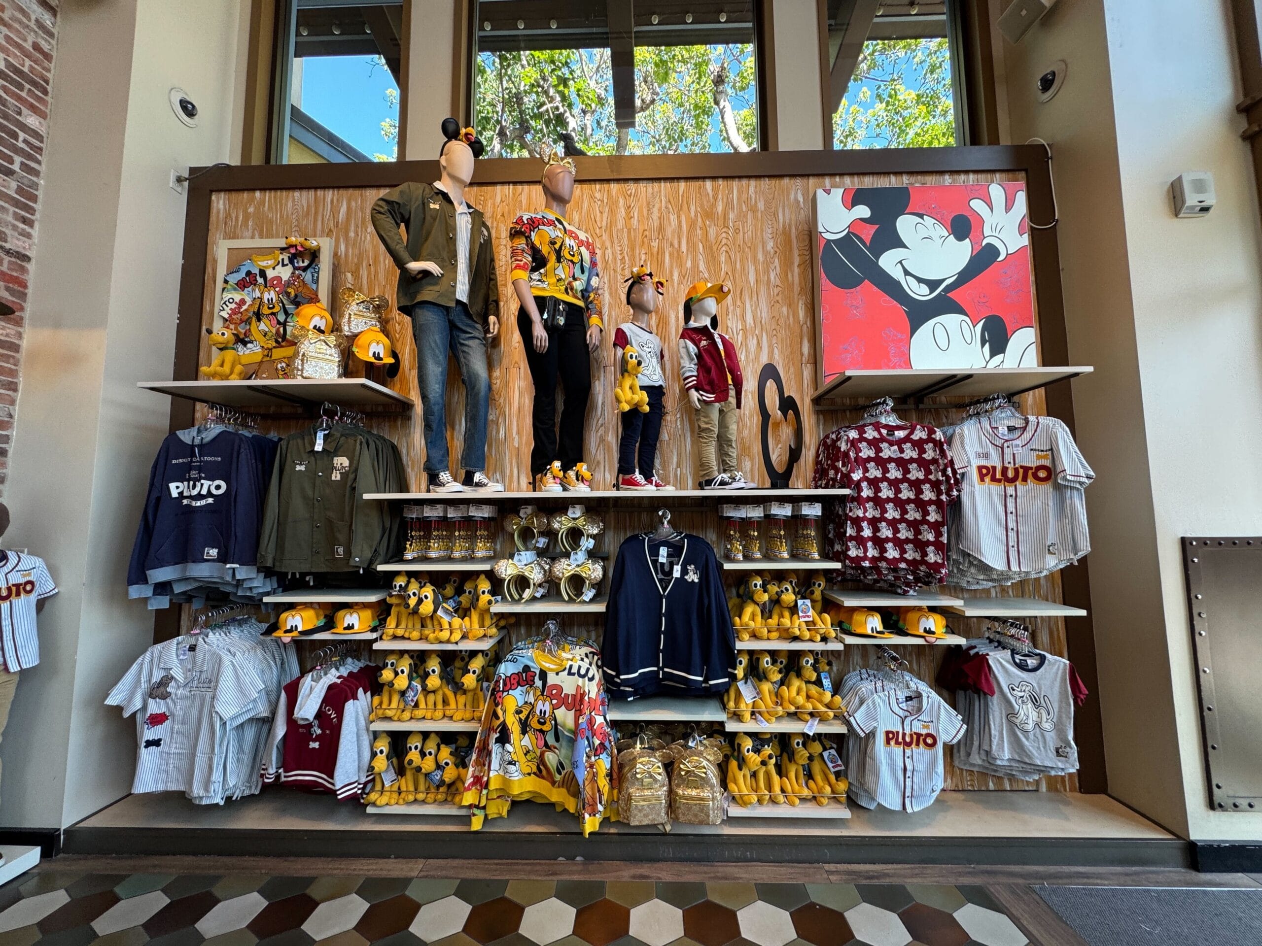 Display of Disney merchandise featuring Pluto plush toys, shirts, jackets, hats, mugs, and a Mickey Mouse wall art.