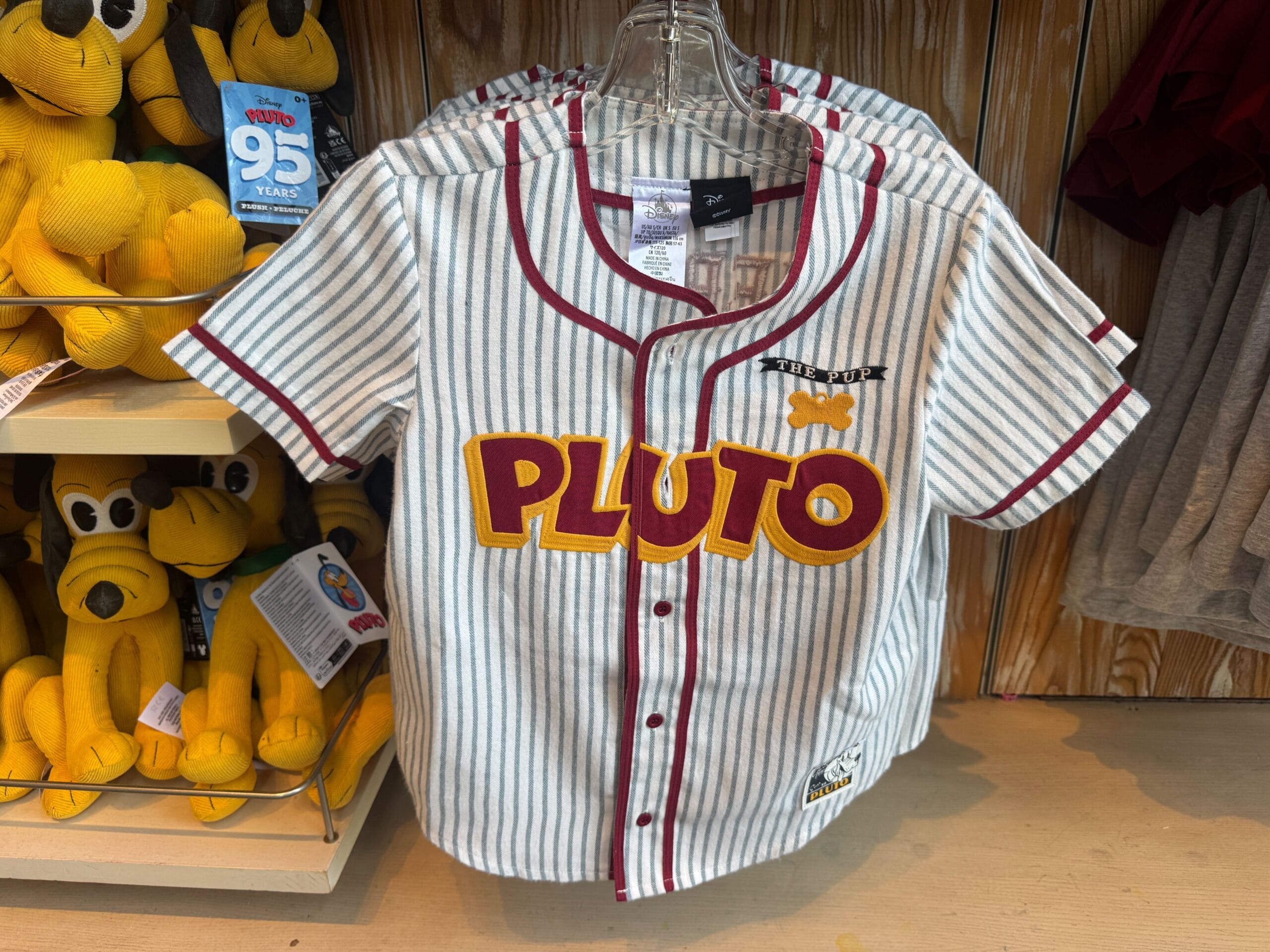 Striped baseball jersey with "PLUTO" in bold letters, Pluto graphic, and Pluto plush toys displayed nearby on a shelf.