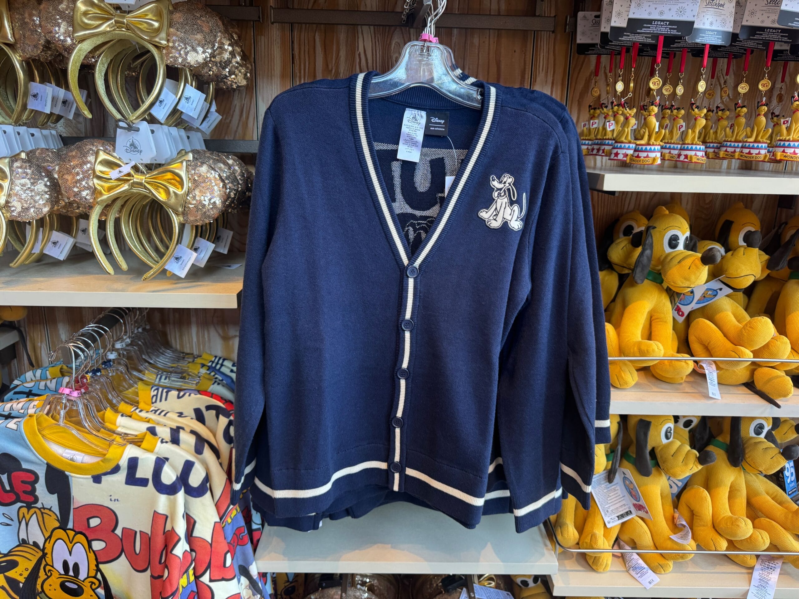 Navy blue cardigan with a Pluto patch displayed on a store rack, surrounded by Pluto plush toys and Disney merchandise.