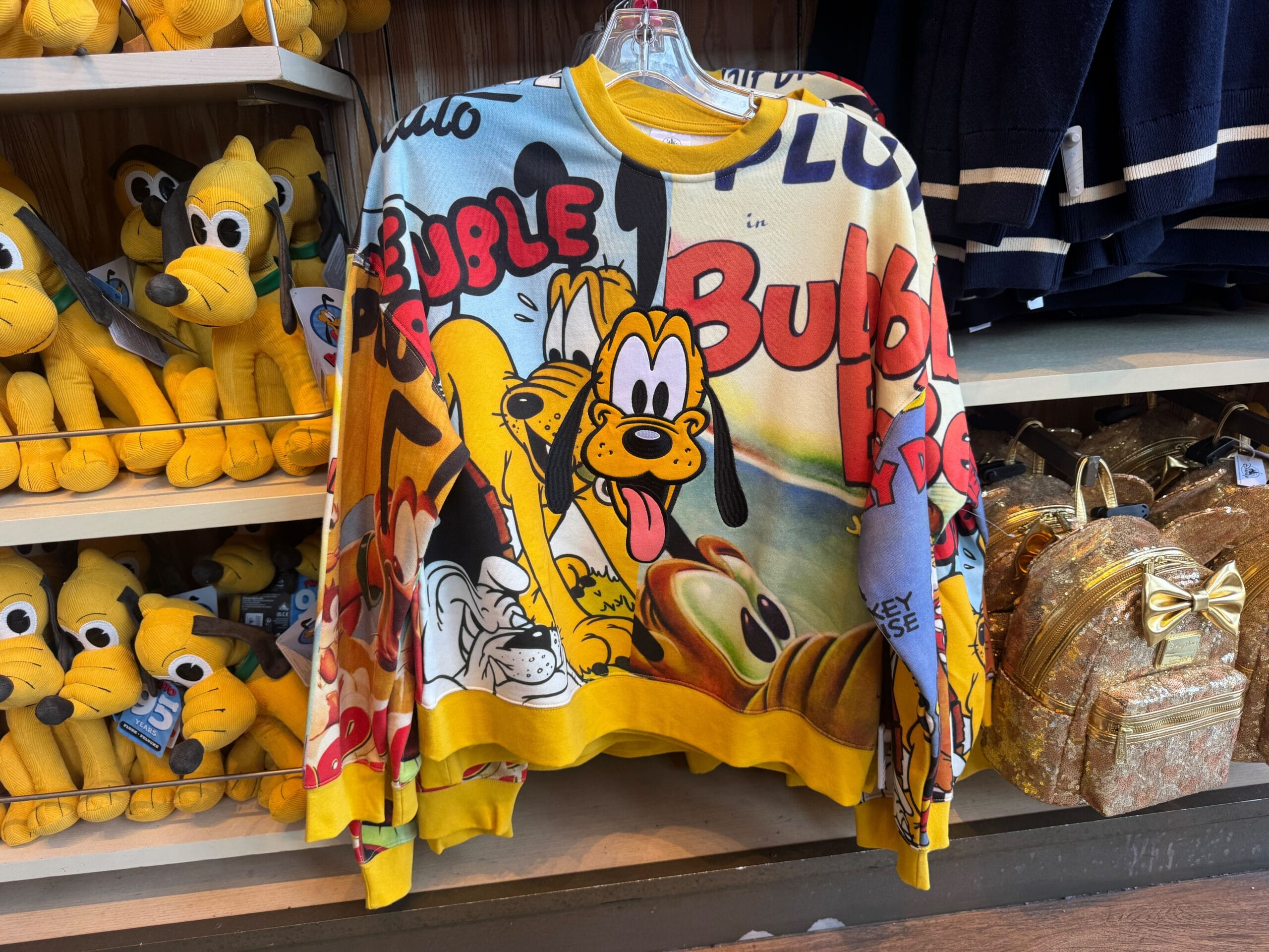 Colorful sweater featuring Pluto and comic graphics, displayed in a store with Pluto plush toys and gold backpacks.