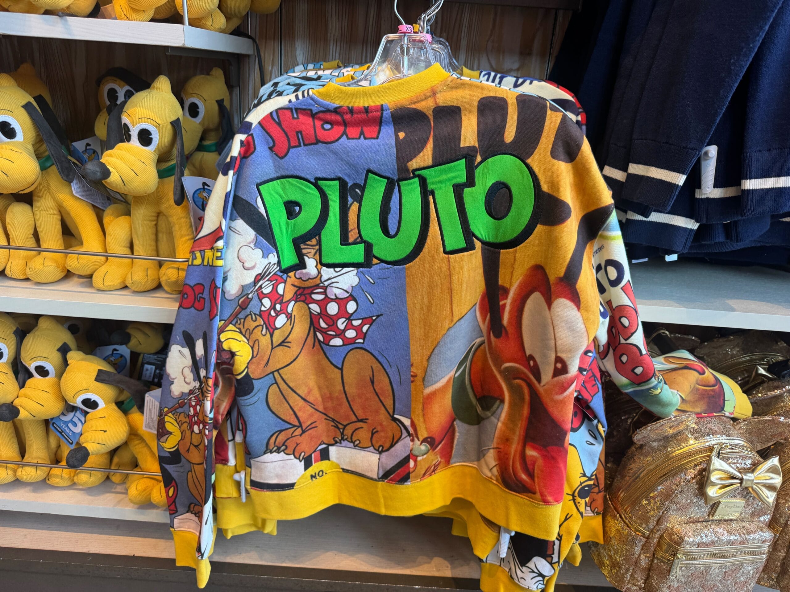 A colorful Pluto-themed sweatshirt hangs on display, surrounded by plush Pluto toys and a gold backpack.