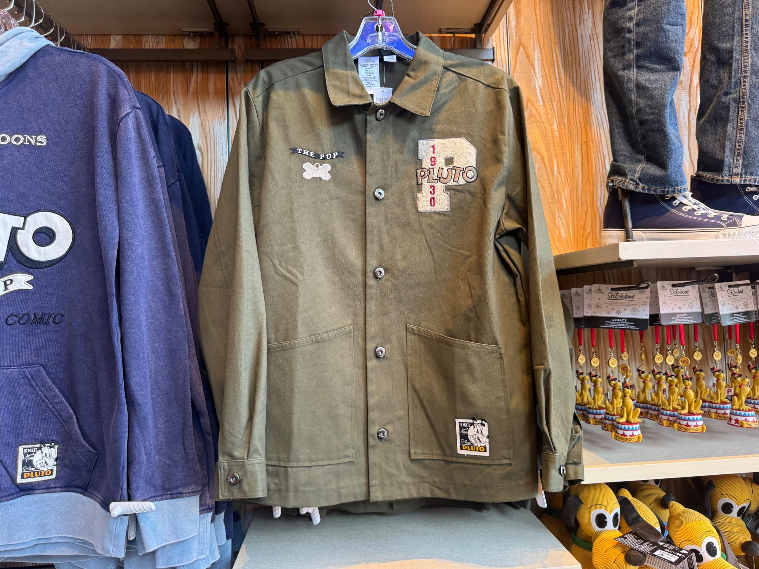 Olive green Pluto jacket with patches hangs on a rack, surrounded by other Disney-themed clothing and merchandise.