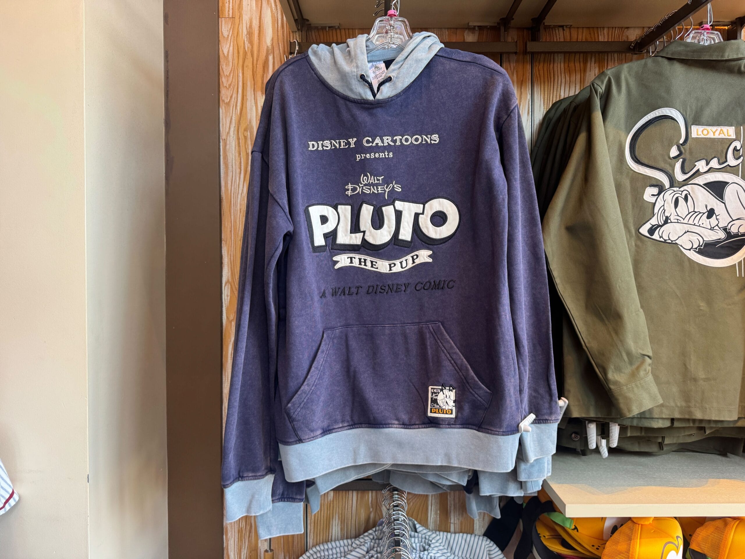 A navy blue Pluto the Pup Disney hoodie hangs on a rack in a store, with another green hoodie beside it.