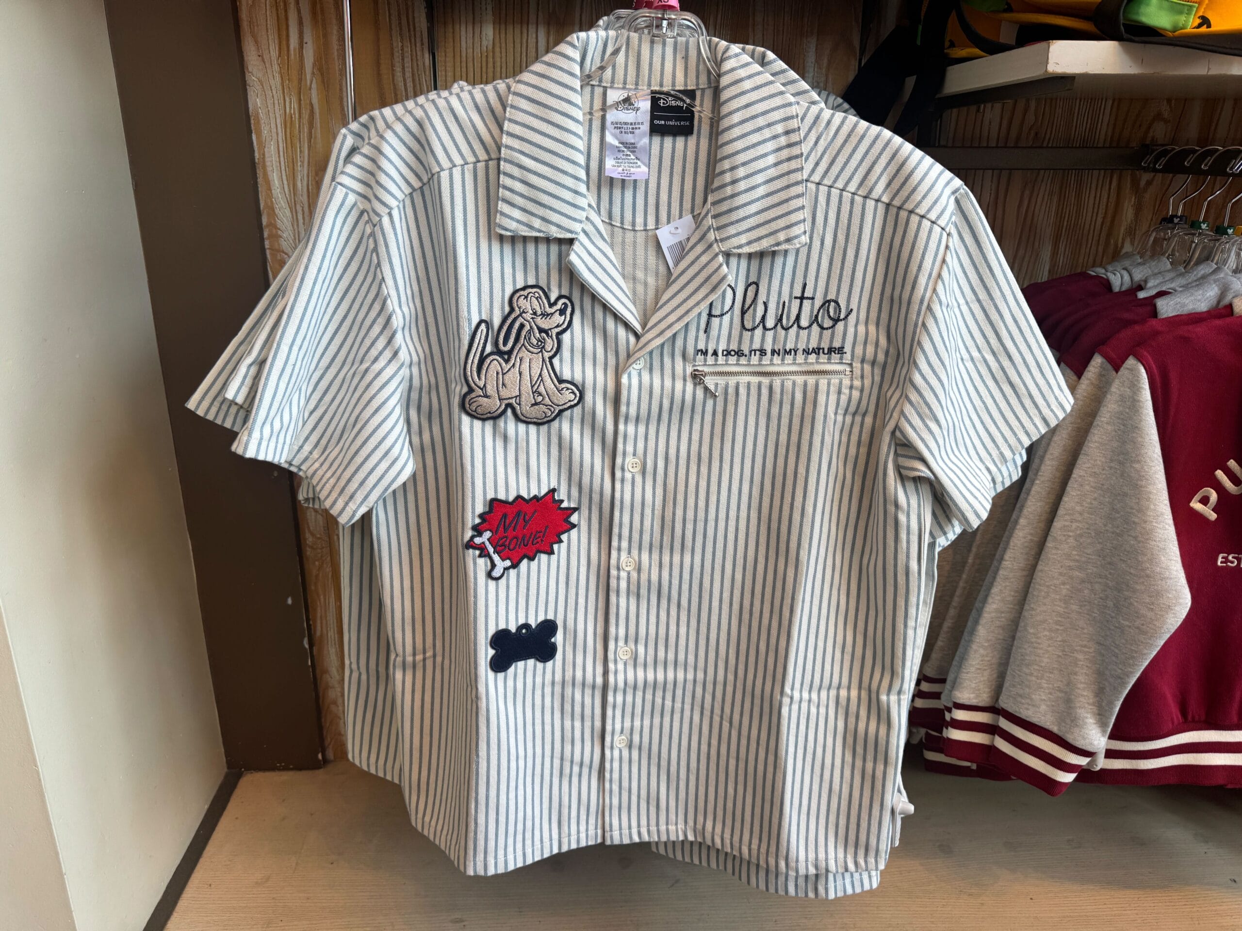Short-sleeve, striped shirt with Pluto dog patches and embroidered "Pluto" text displayed on a hanger in a store.