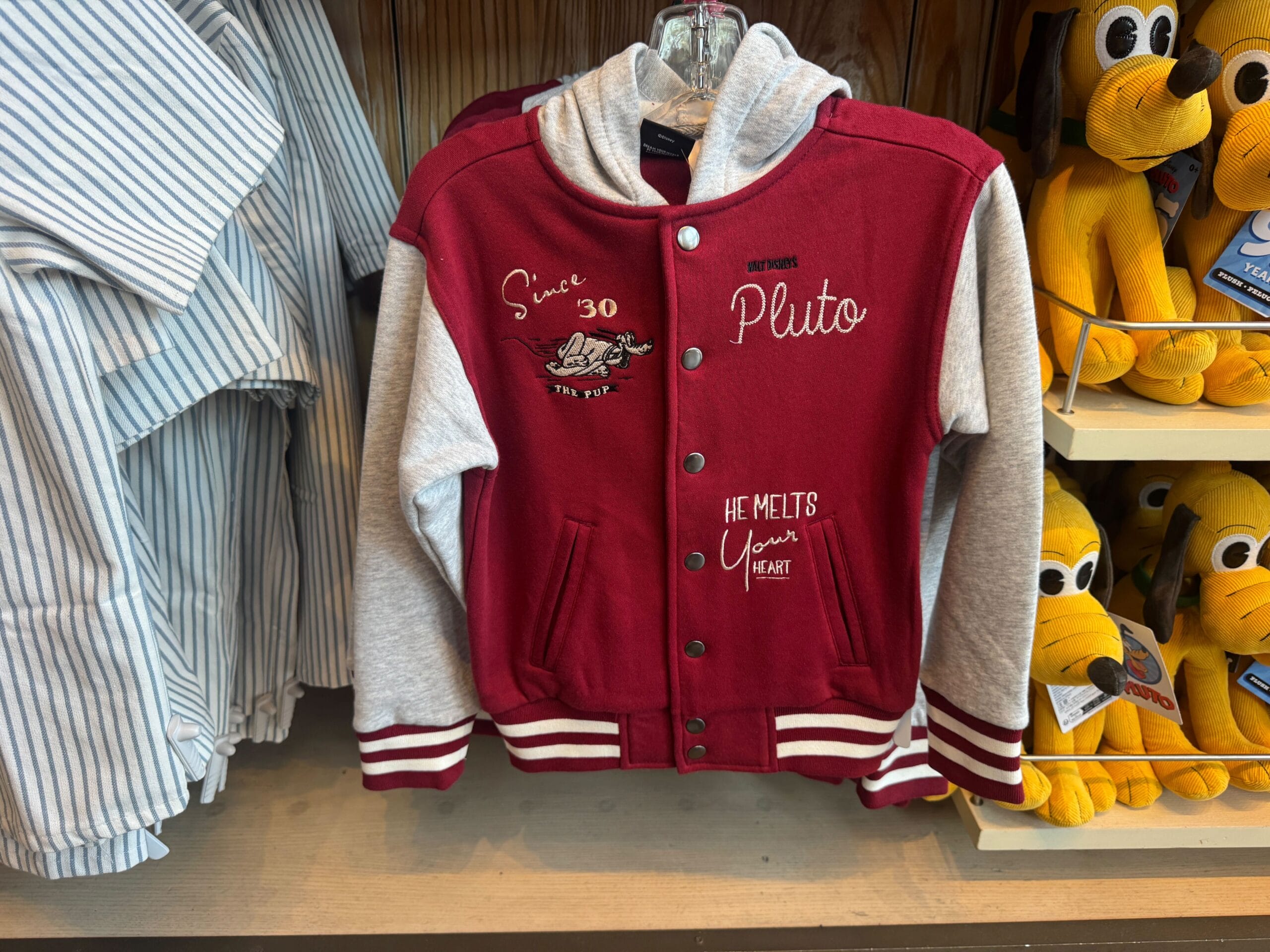 Red and gray Pluto-themed jacket with text and graphics, hanging on a rack next to plush Pluto toys and striped shirts.