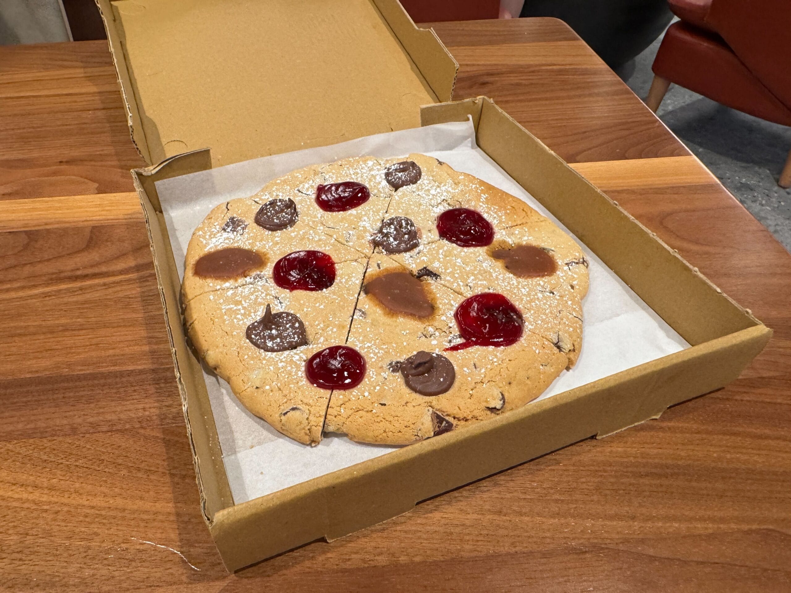 A large Sketch Pad Cookie topped with chocolate and red jelly sits in a pizza box on a wooden table.