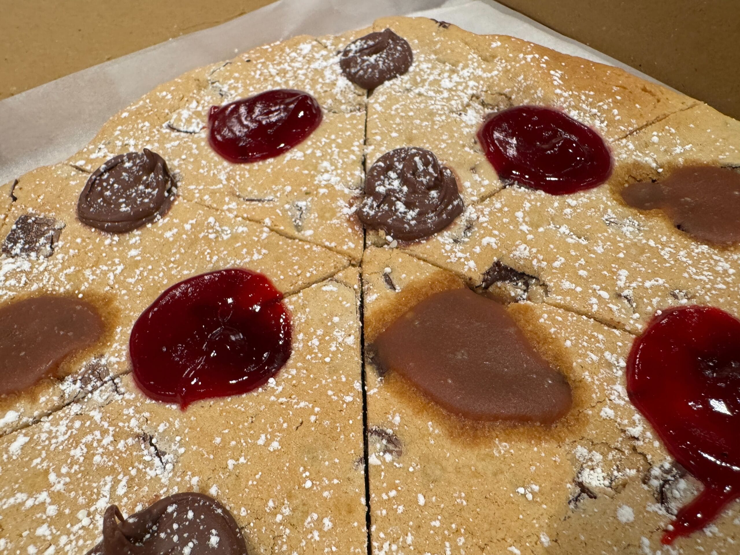 The Sketch Pad Cookie pizza is topped with powdered sugar, chocolate dollops, and raspberry jam, sliced into eight pieces.