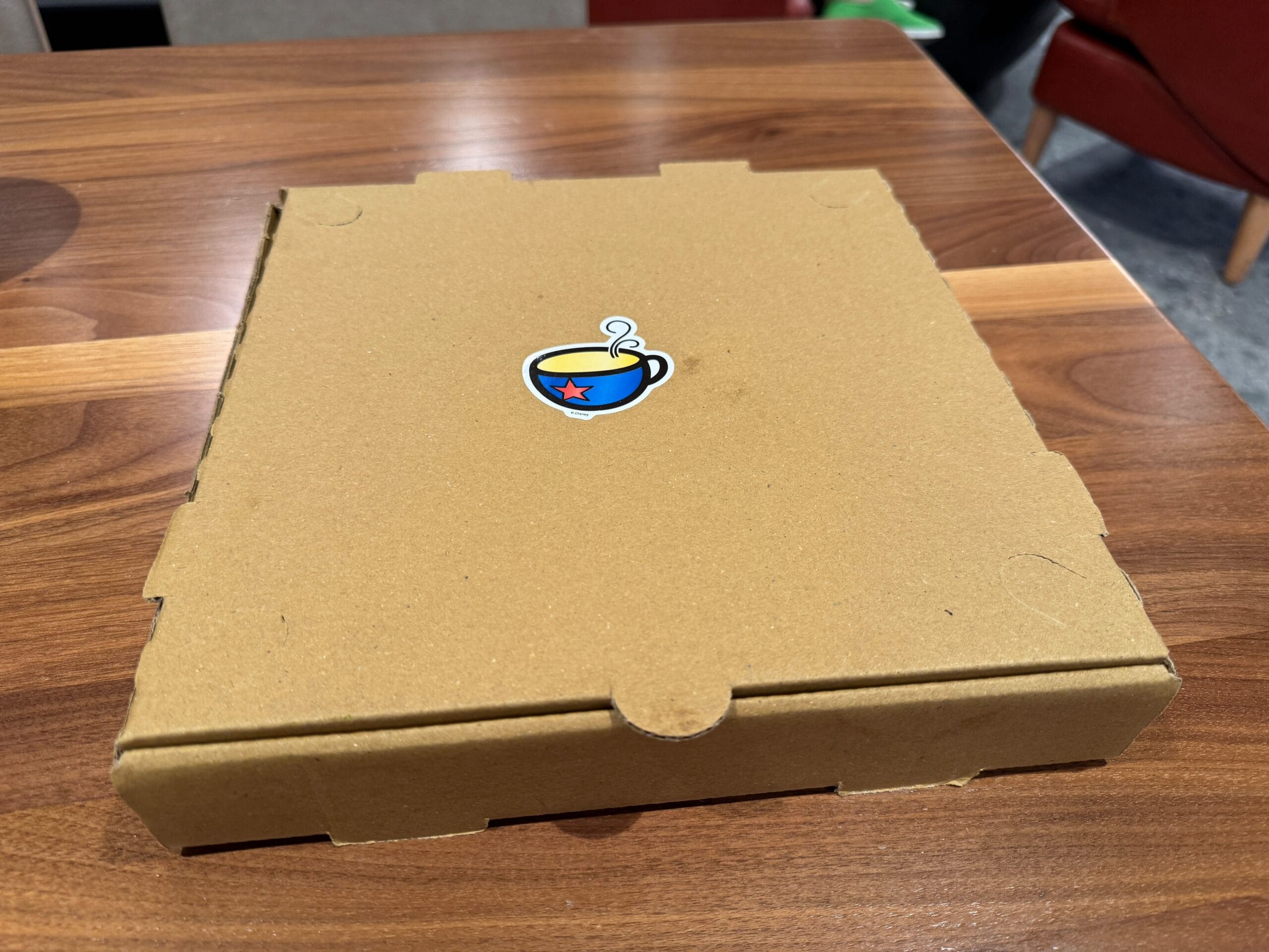 A closed brown pizza box with a Sketch Pad Cookie sticker sits on a wooden table.