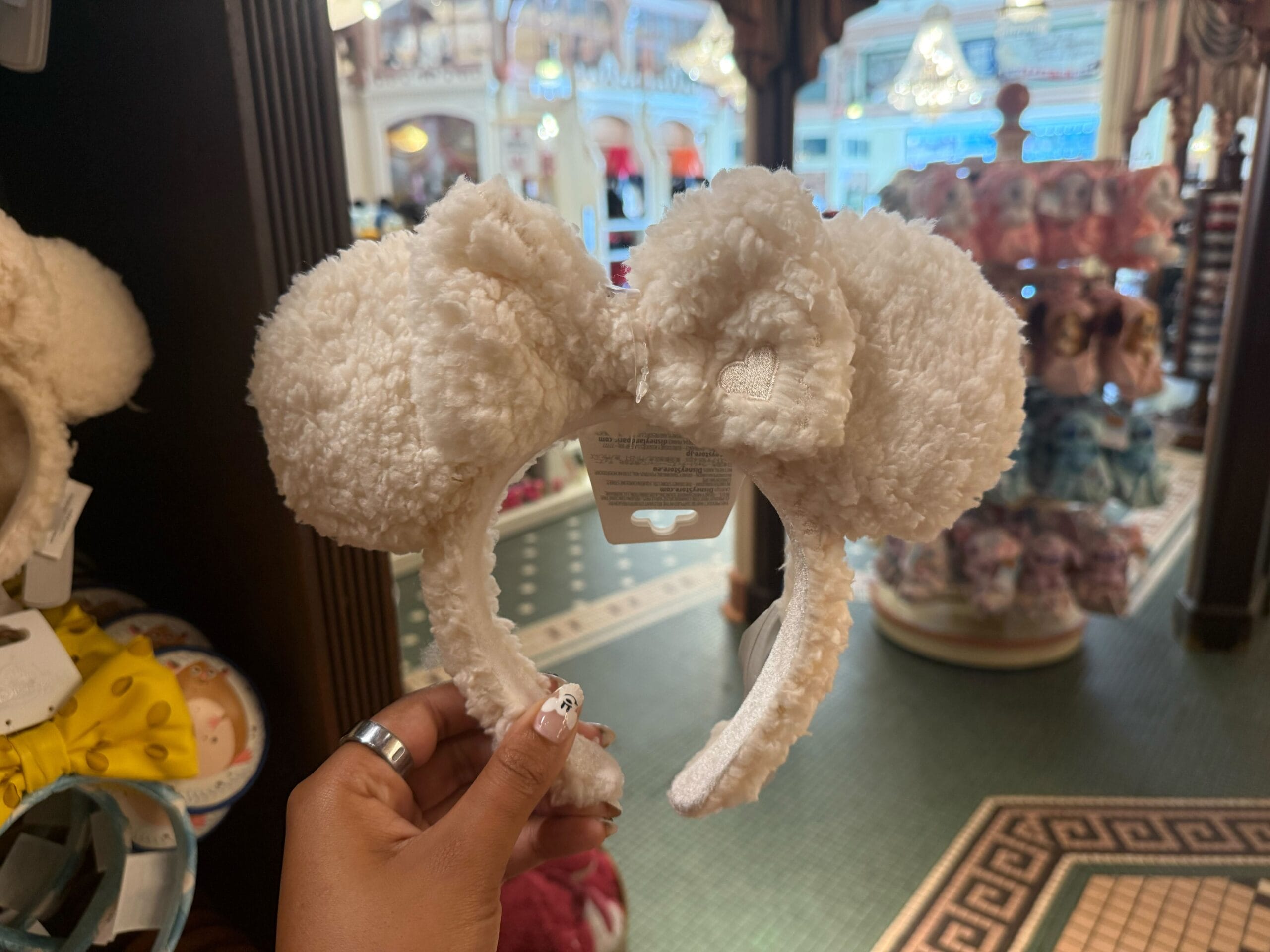 A hand holds a fluffy white Minnie Mouse ear headband with a bow in a store filled with plush toys.