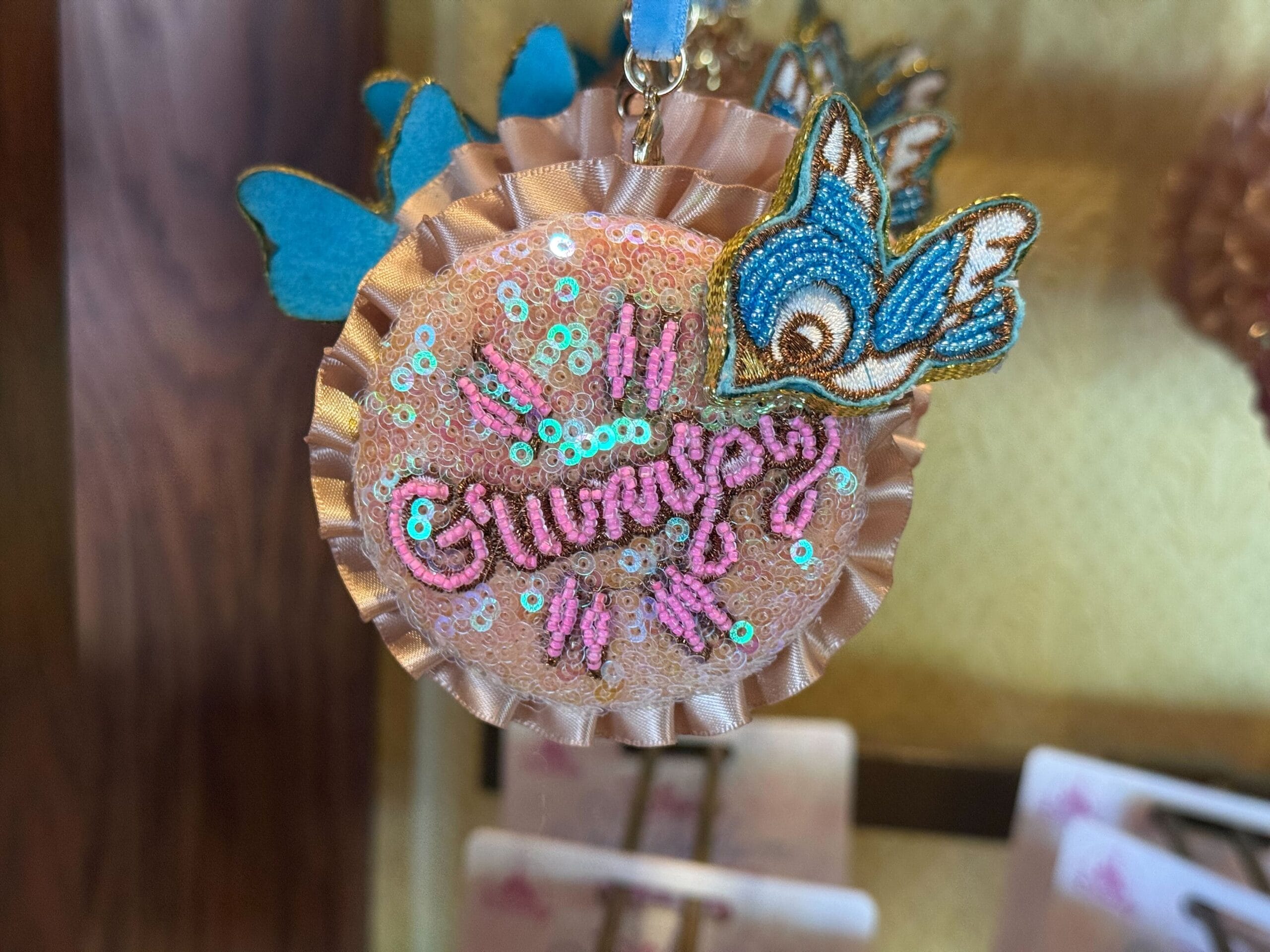 A pink and gold sequined badge from the Ornament Collection with "Grumpy," a blue bird, and blue butterflies.