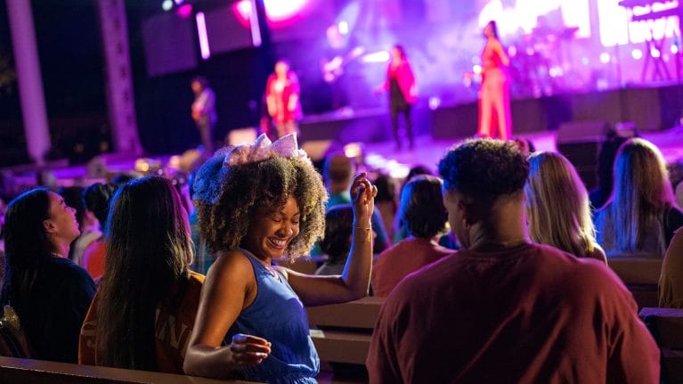 People enjoy a live music concert, with a smiling woman dancing in the foreground and colorful stage lights in the background.