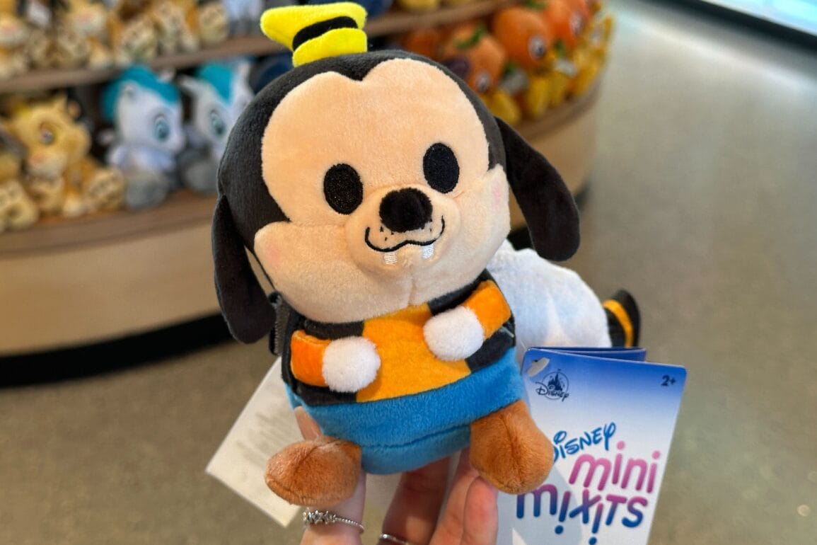 A hand holds a plush Goofy toy in a store at Walt Disney World, with other Disney plushies blurred in the background.