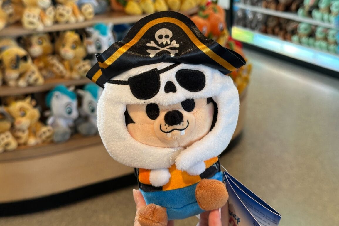 A hand holds a plush pirate toy with a skull hat and eye patch at a Walt Disney World store filled with plush toys.