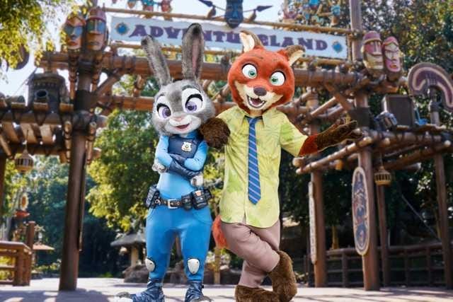 Two people in Judy Hopps and Nick Wilde costumes stand smiling in front of an Adventureland entrance at a theme park.