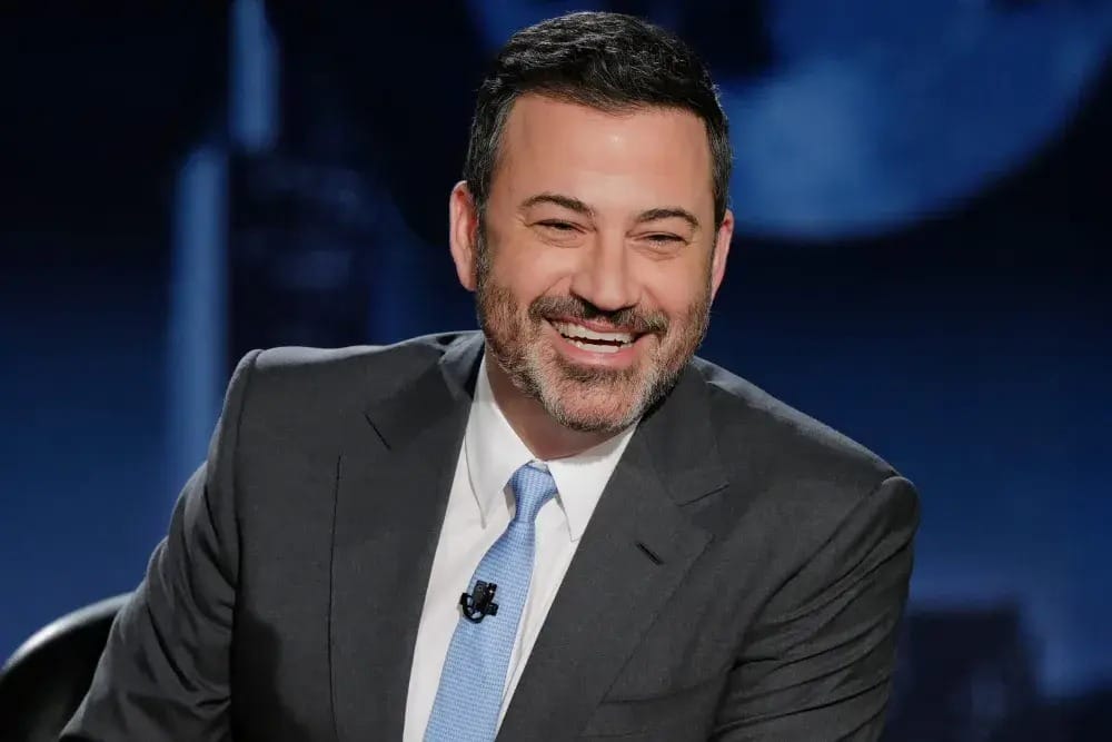 A man in a suit and tie smiles while seated in a studio, resembling a Disney live show set with a dark background.