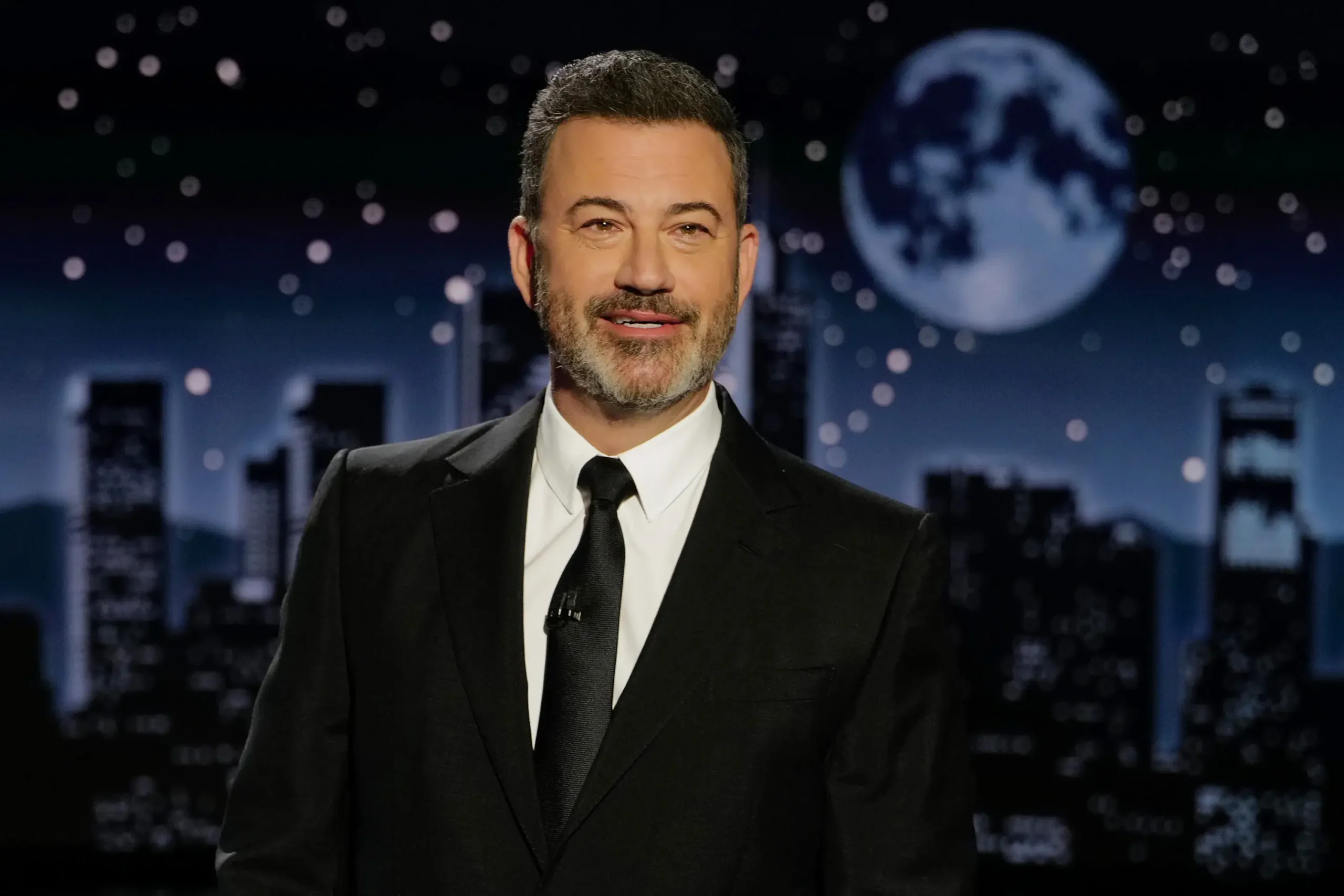 A man in a black suit smiles before a city skyline at night, as seen on Jimmy Kimmel Live on ABC.