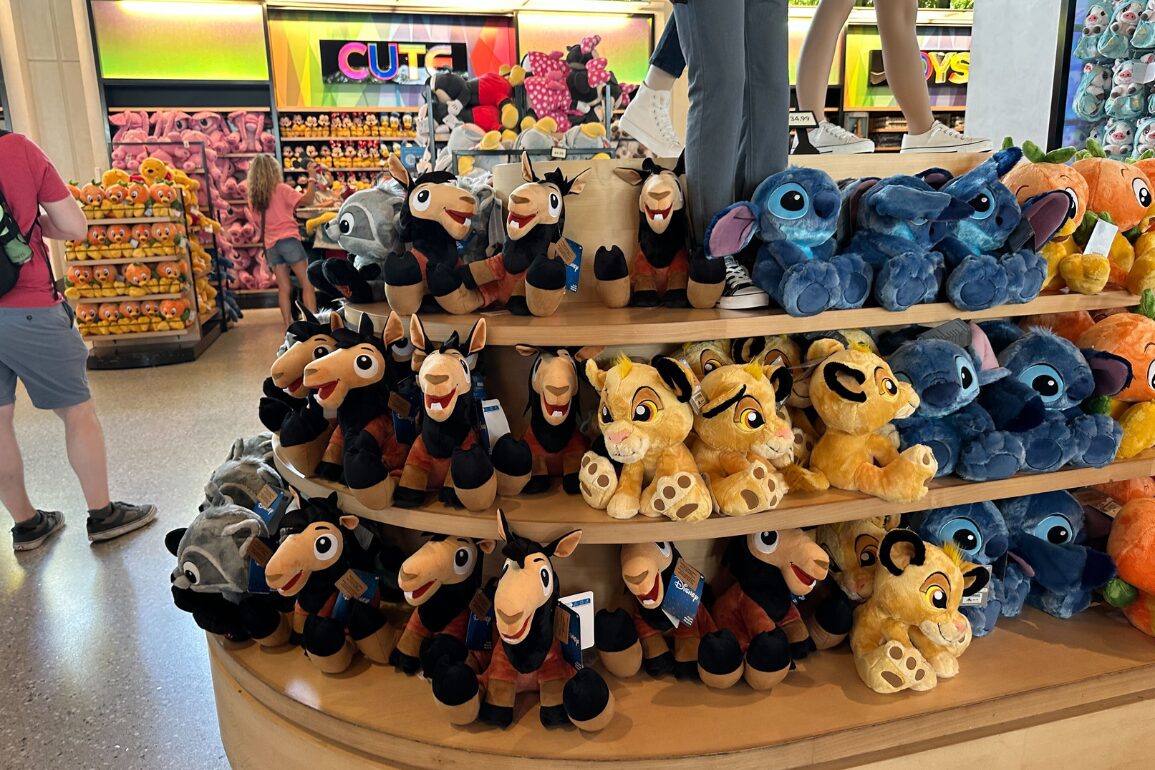 Plush toys from Disney movies displayed on tiered shelves in a store, with colorful signs and shoppers in the background.