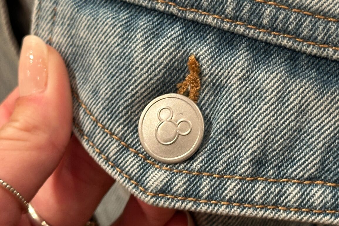 A hand holds a Mickey Denim Jacket with a silver Disney button, near a small piece of brown debris.