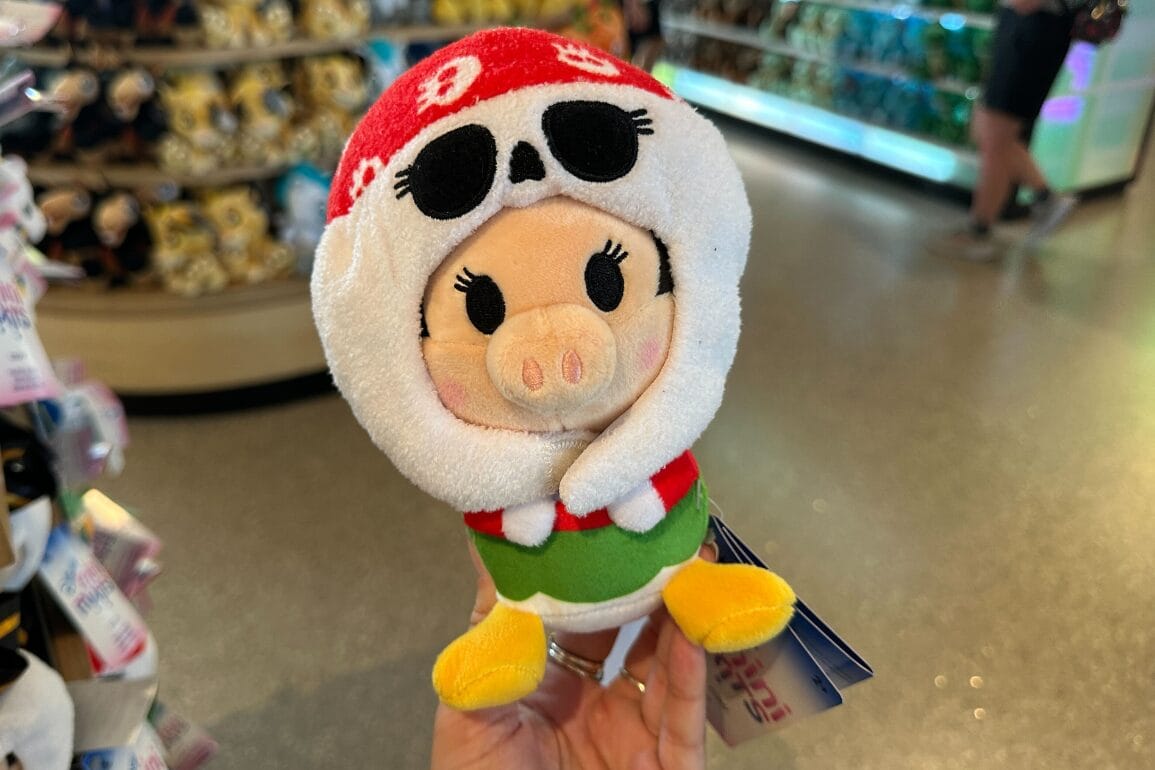 A hand holds a plush toy pig in a red mask with skull, green top, and yellow shoes at Walt Disney World store.