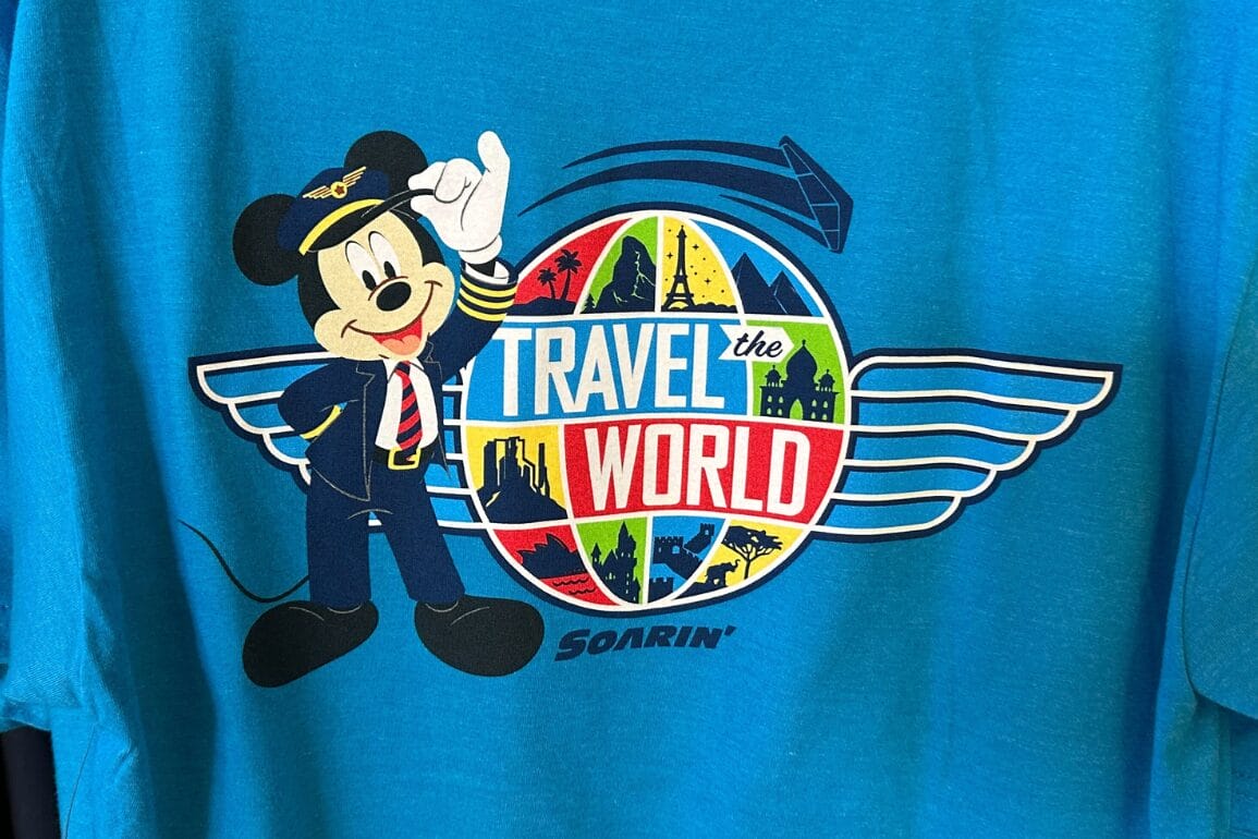 Mickey Mouse in a pilot uniform stands by a "Travel the World" globe, Soarin' T-Shirt style, on a blue EPCOT shirt.