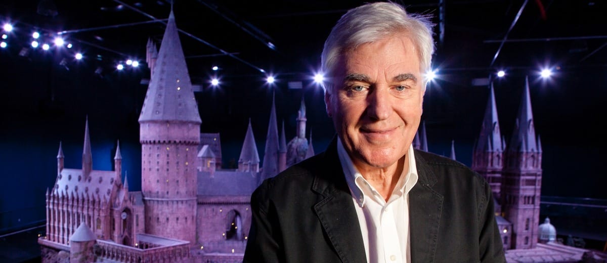 An older man in a suit smiles by a castle model, echoing Harry Potter’s magic—perhaps a nod to production designer Stuart Craig.