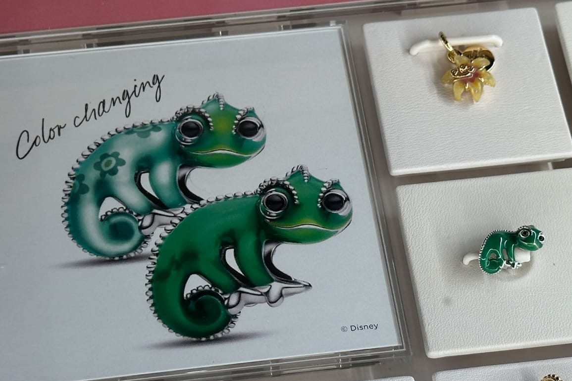 Display case with charms from Pandora's Tangled collection: a yellow sundrop flower charm on top and a green Pascal chameleon charm below, with a descriptive sign on the left showing the chameleon charm's color-changing properties.