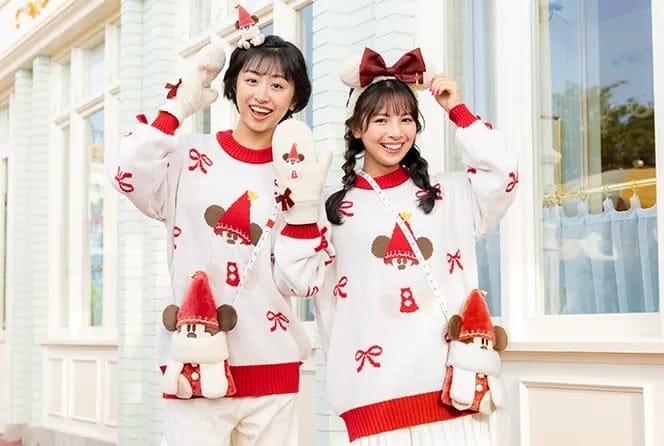 Two smiling people in matching festive sweaters with cartoon mouse designs, posing outside Tokyo Disneyland.