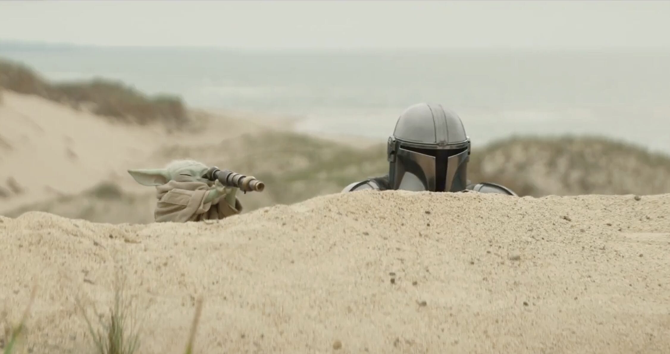 Grogu and the Mandalorian hide behind a sand dune on a beach, with the ocean visible in the background.