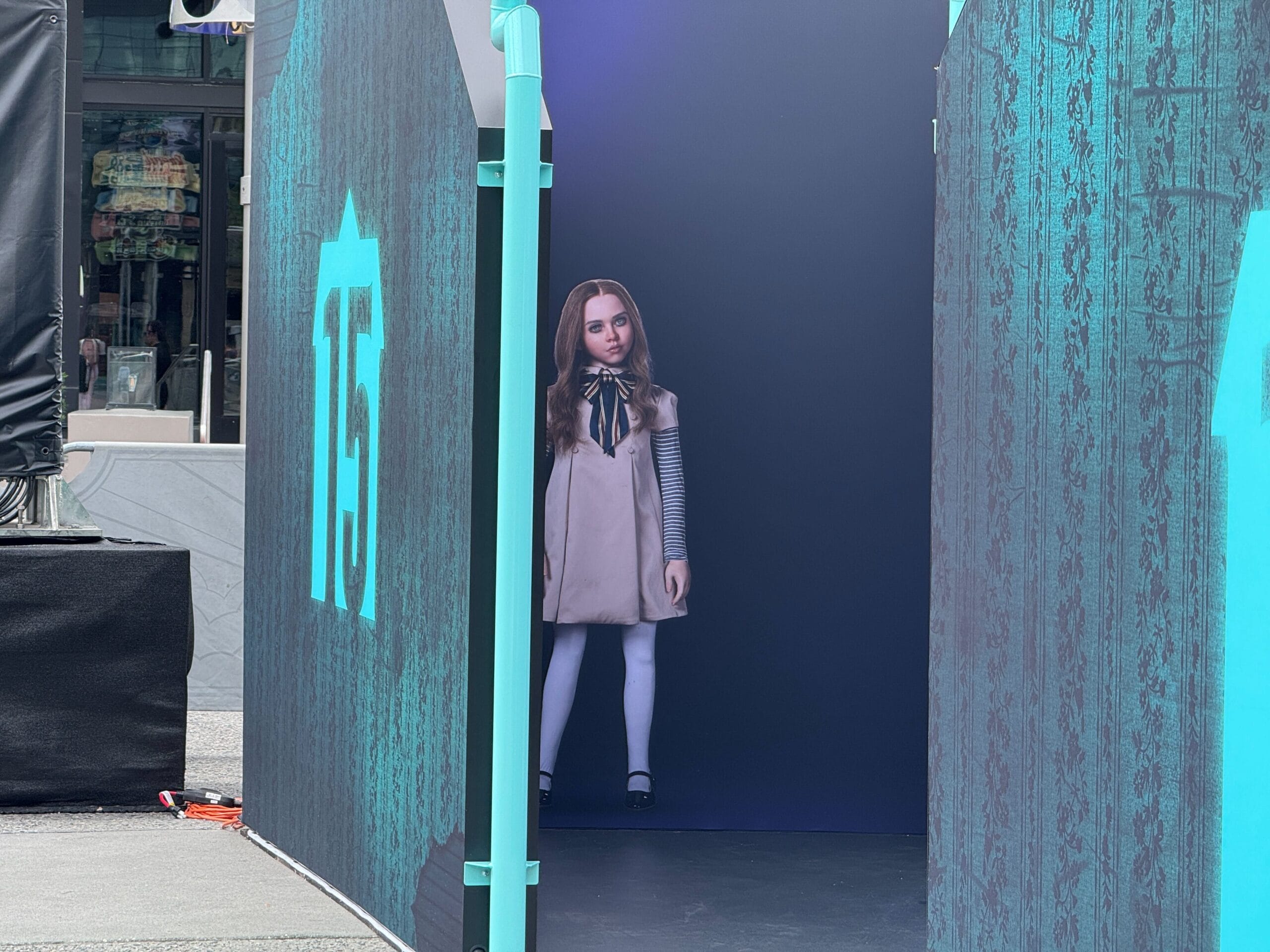 A life-size cutout of a doll-like girl stands inside a blue, partially enclosed display at an outdoor event.