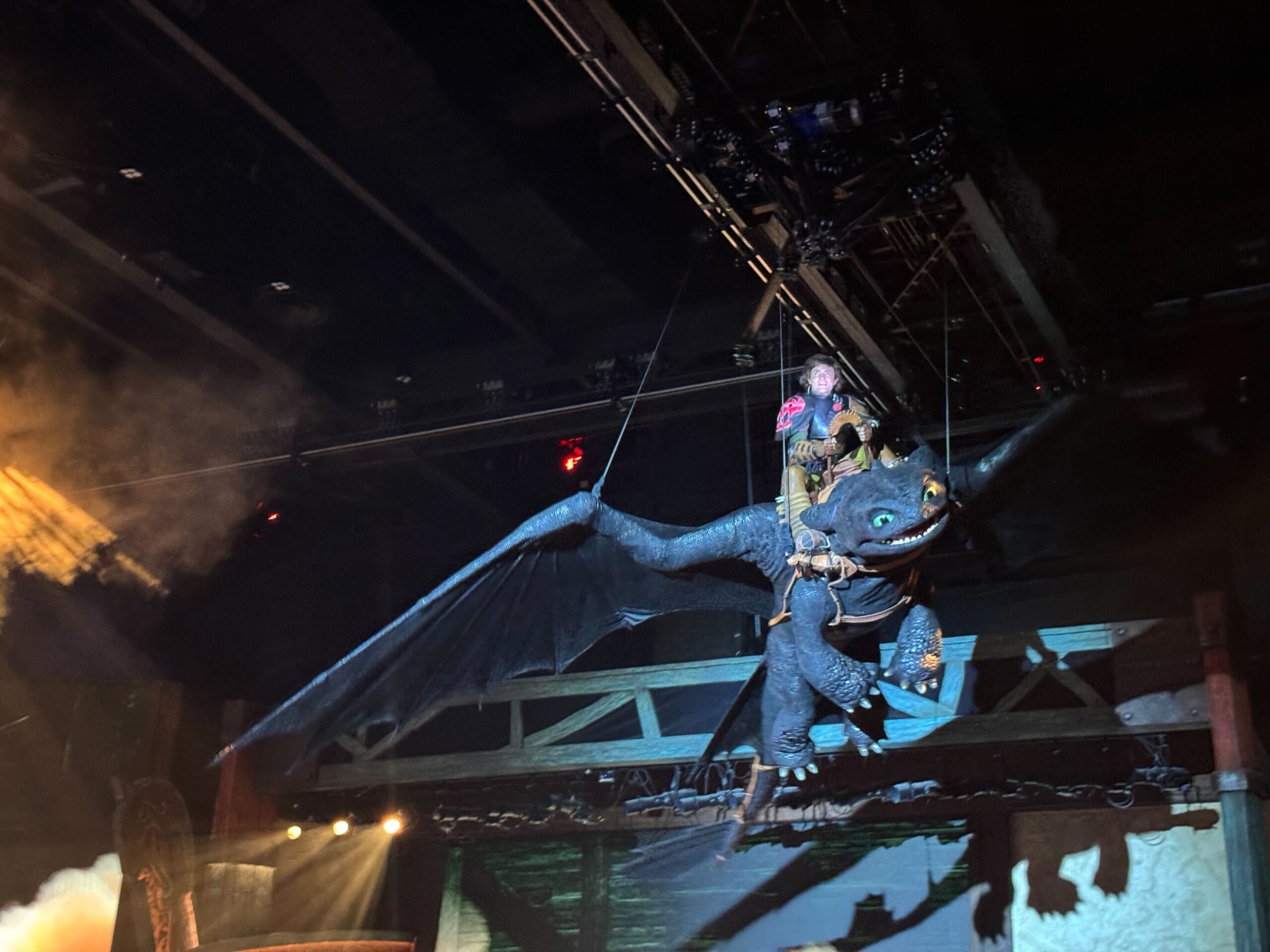 Performer rides a large, black Toothless dragon puppet suspended in the air during an epic stage show.