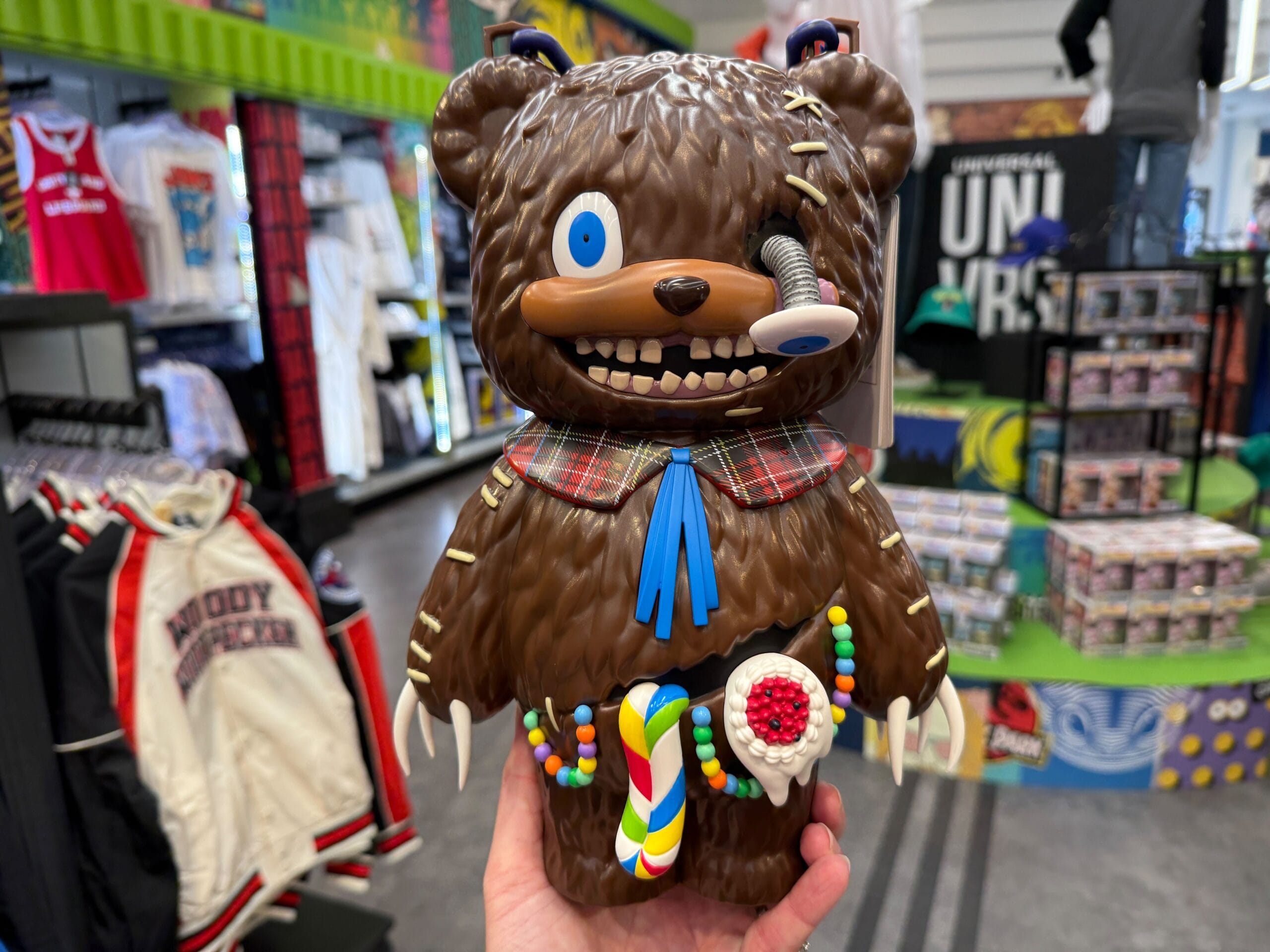 A hand holds a quirky bear figurine with a spring eye, plaid collar, and colorful candy decorations in a store.