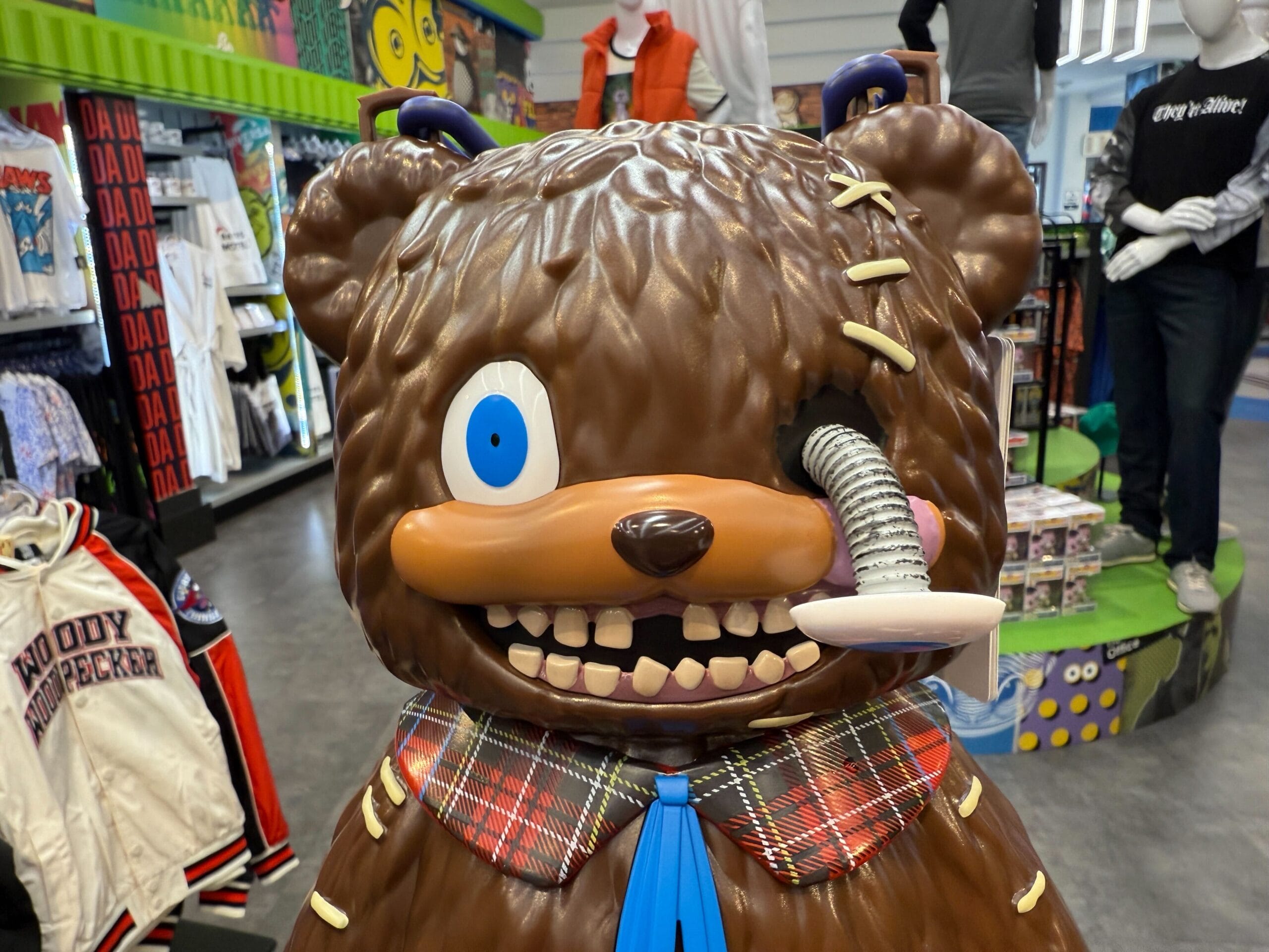 A cartoonish bear statue with a mismatched eye, large teeth, and a metal tube in its cheek stands in a clothing store.