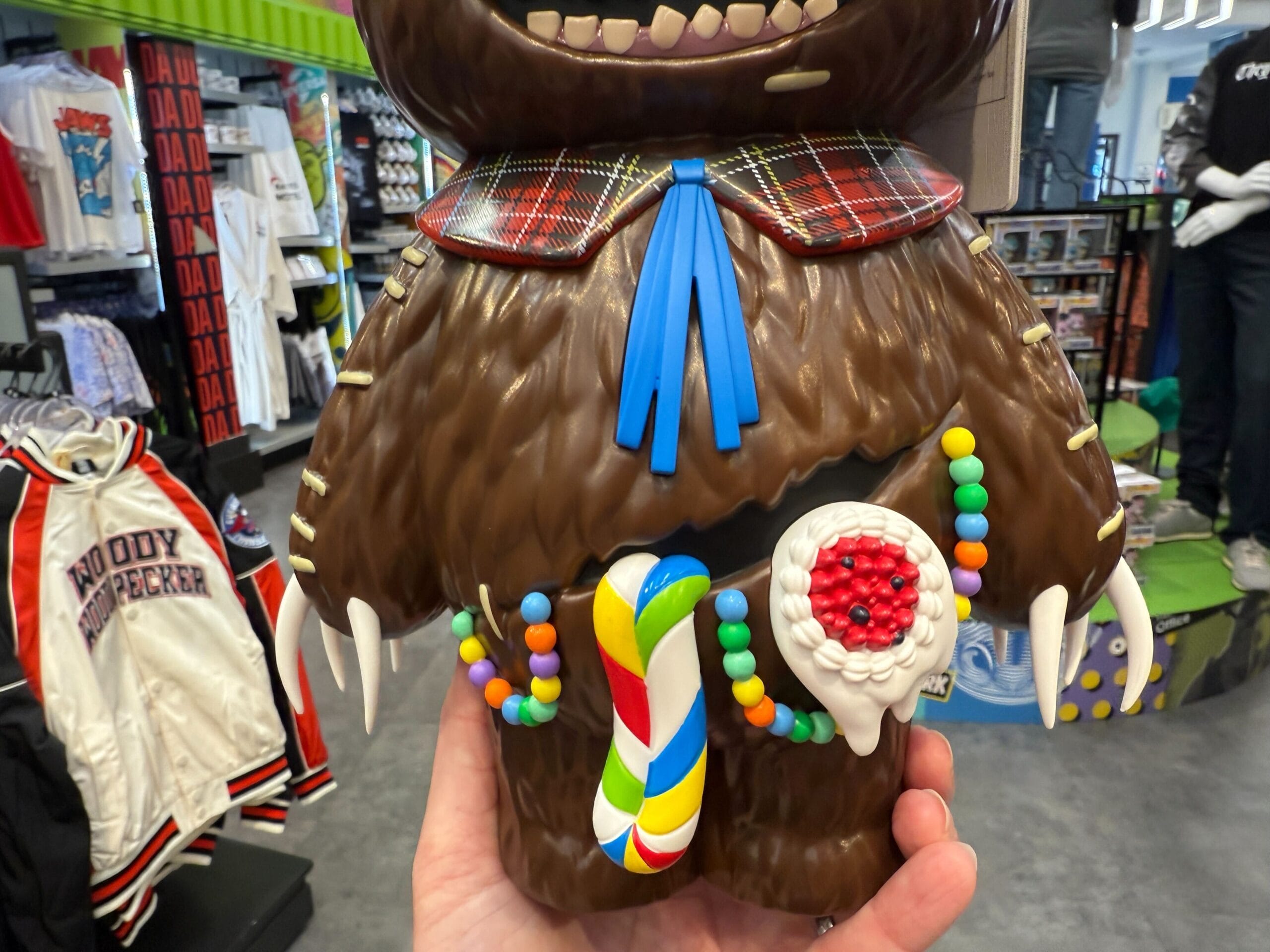 A hand holds a furry toy with a candy necklace, colorful lollipop, and plaid collar in a clothing store.