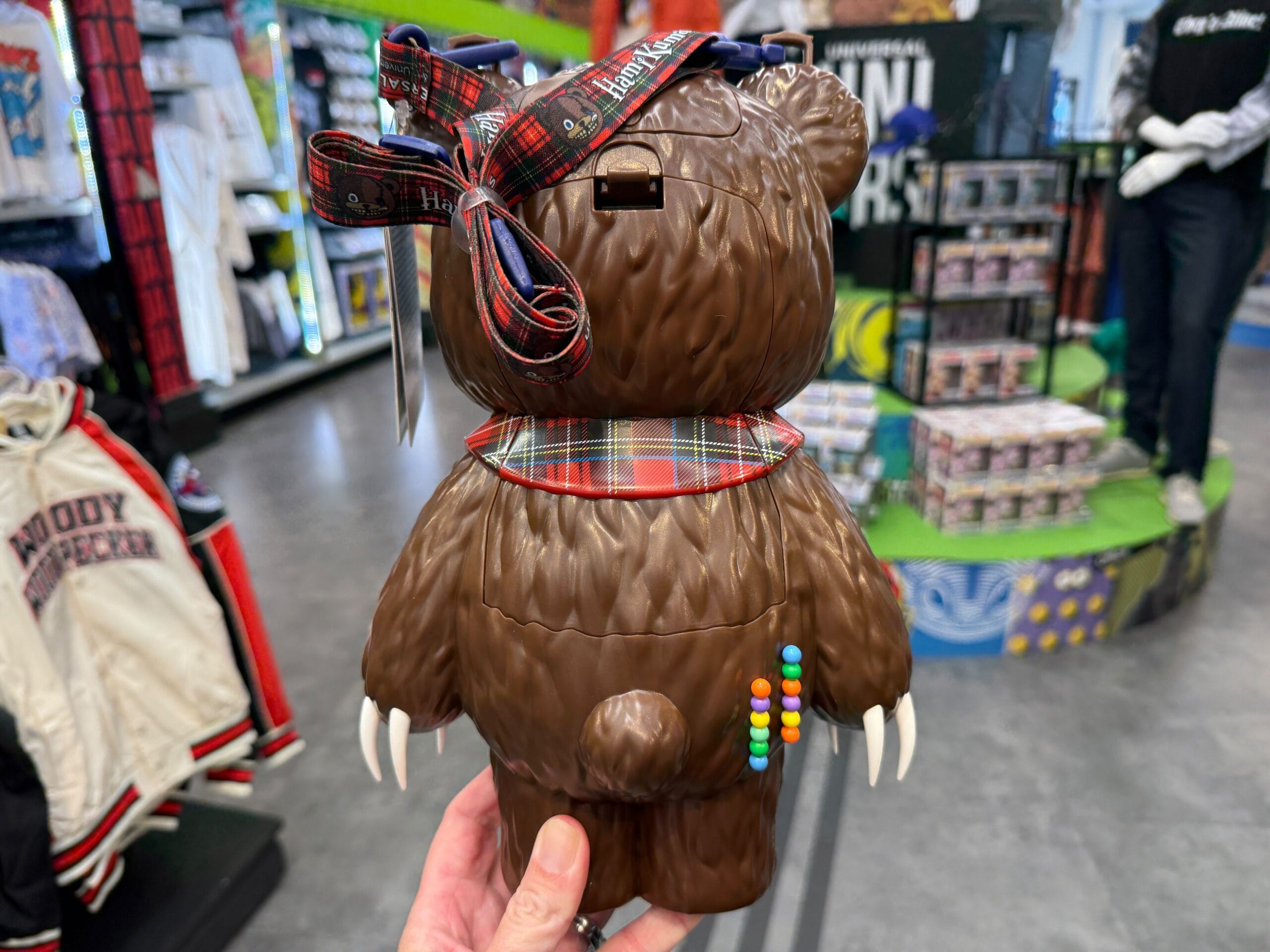 A hand holds a bear-shaped container with claws, a plaid bow, and colorful candy pieces on its side in a store.
