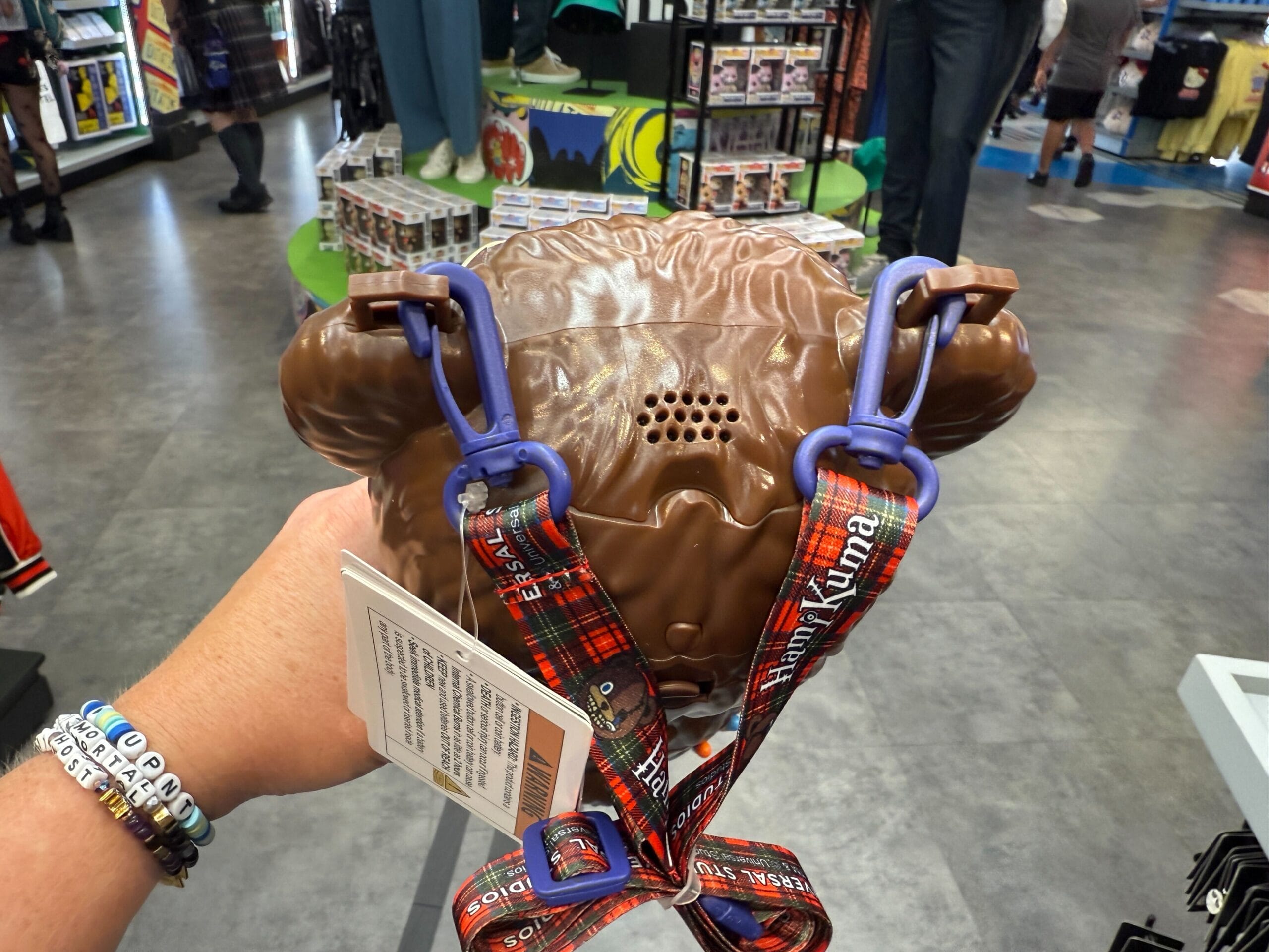 A hand holds a brown Ewok-shaped bag with a plaid strap inside a colorful store.