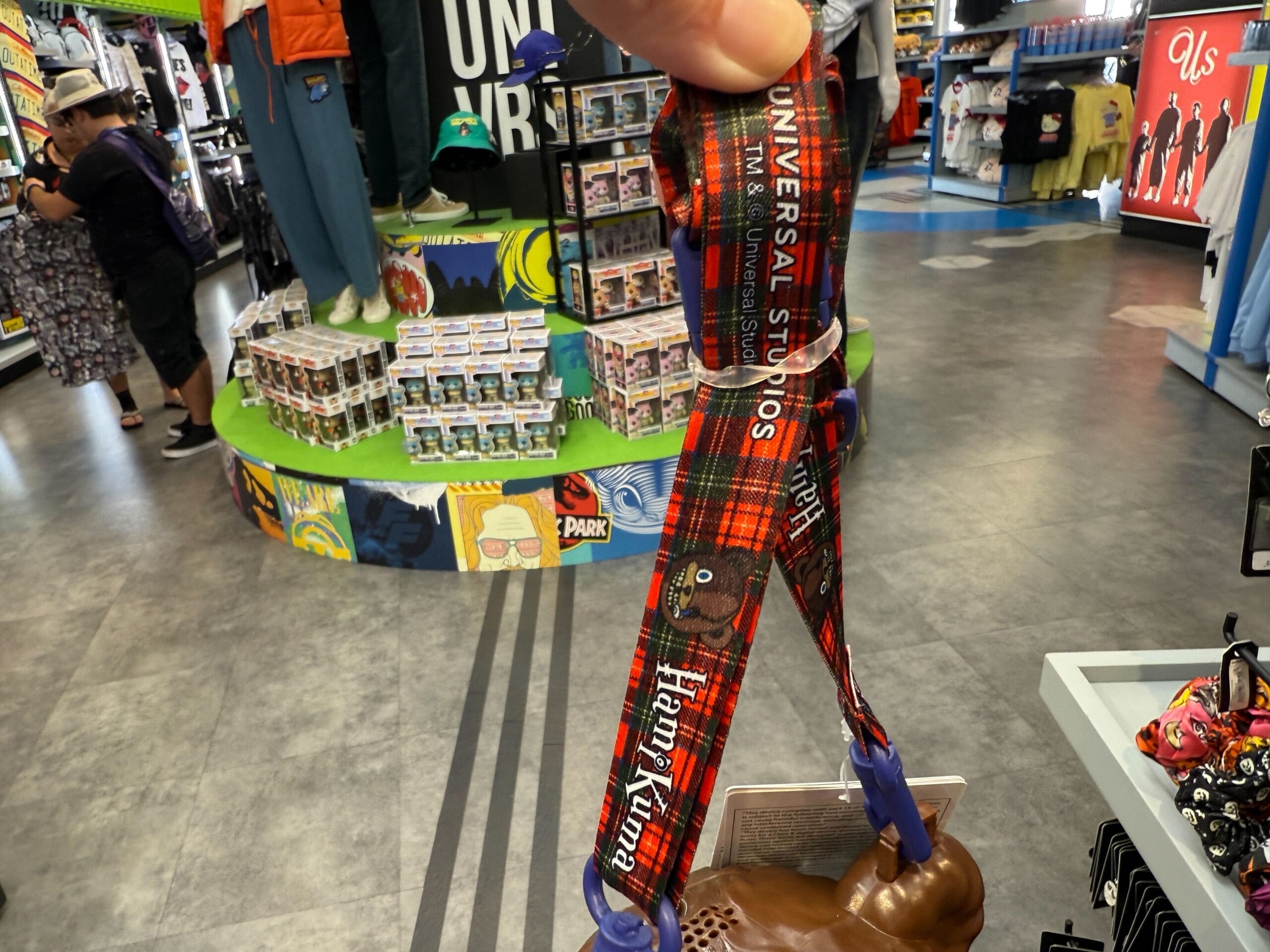 Hand holding a red Universal Studios lanyard in a souvenir shop with merchandise and Funko Pop figures on display.