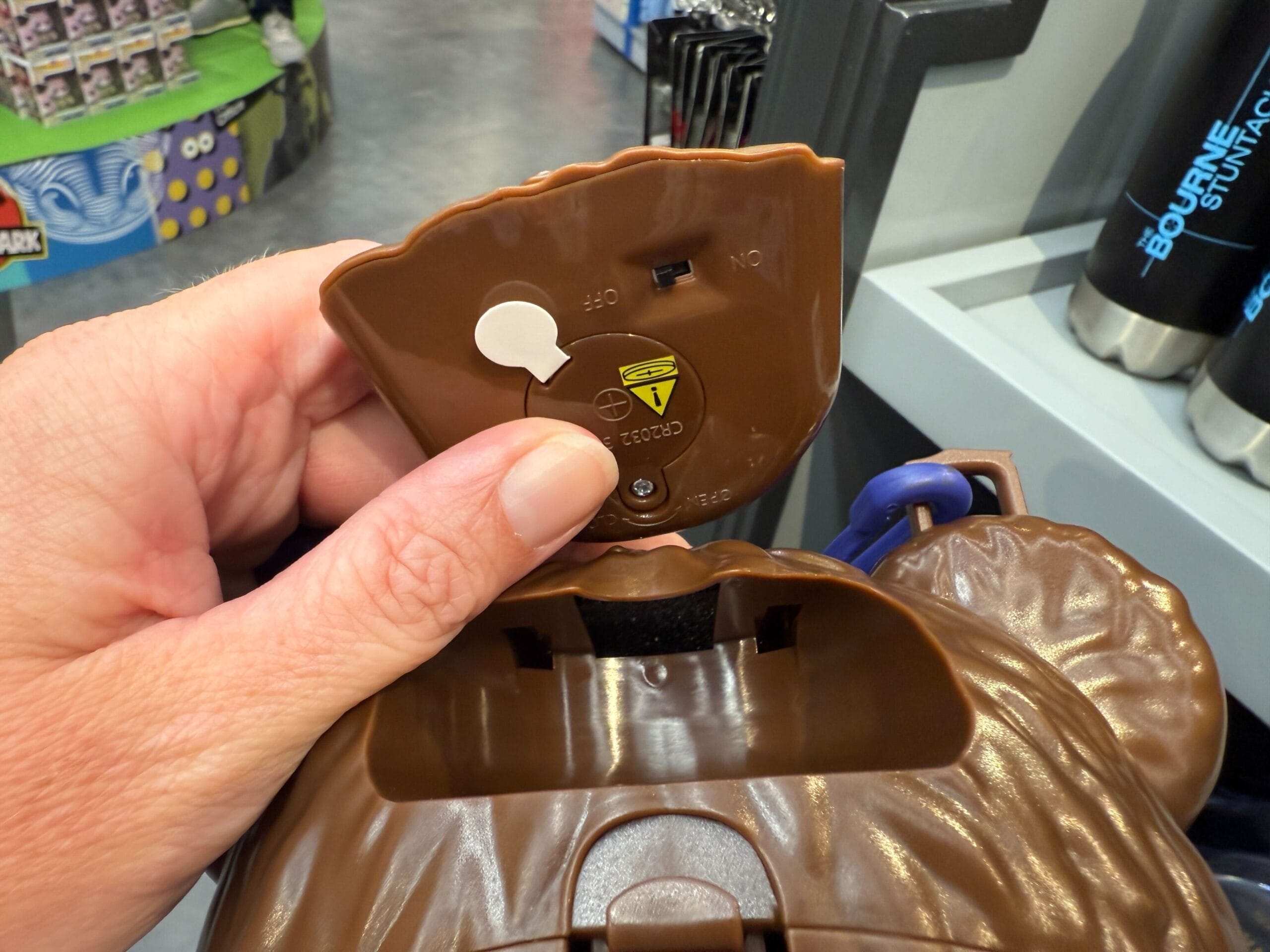 A hand opens a brown plastic compartment showing a battery slot and warning label inside.