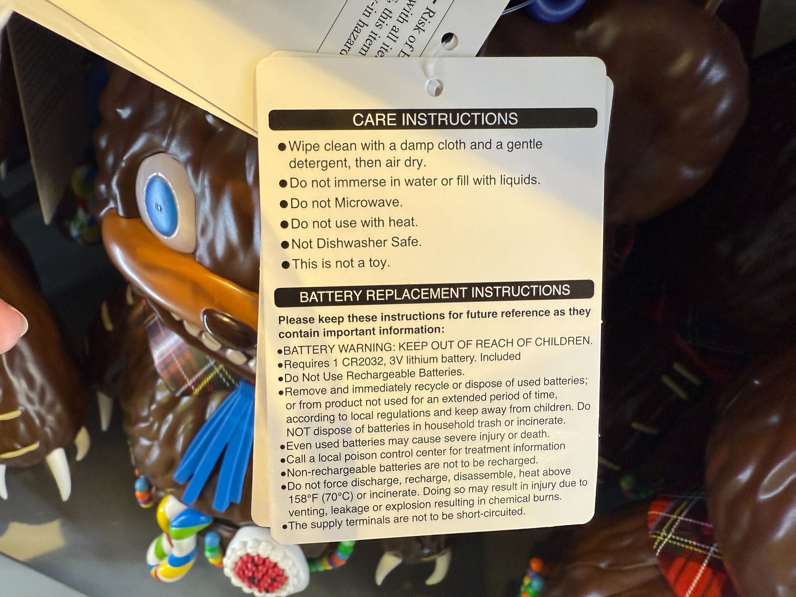 Tag with care and battery instructions hangs in front of a brown toy with orange eyes and colorful decorations.