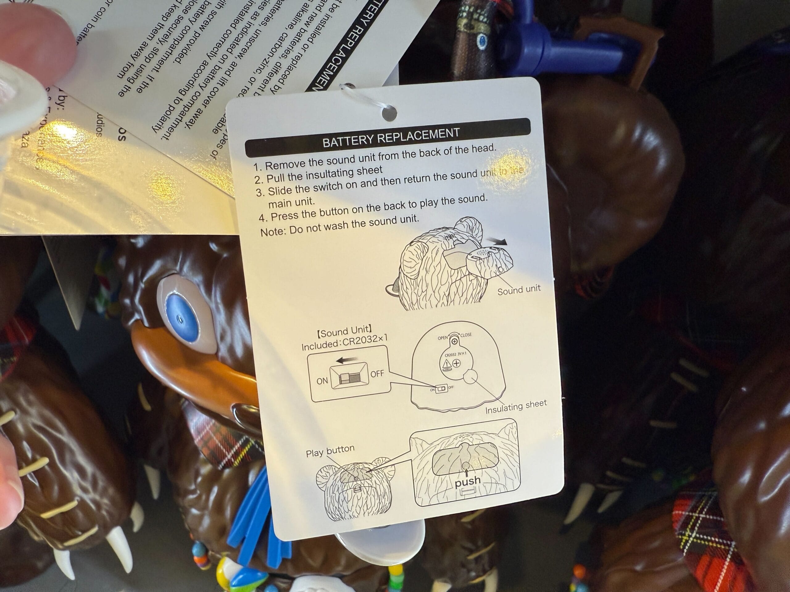 Instruction tag showing battery replacement steps with diagrams for a sound unit attached to a brown, furry toy.