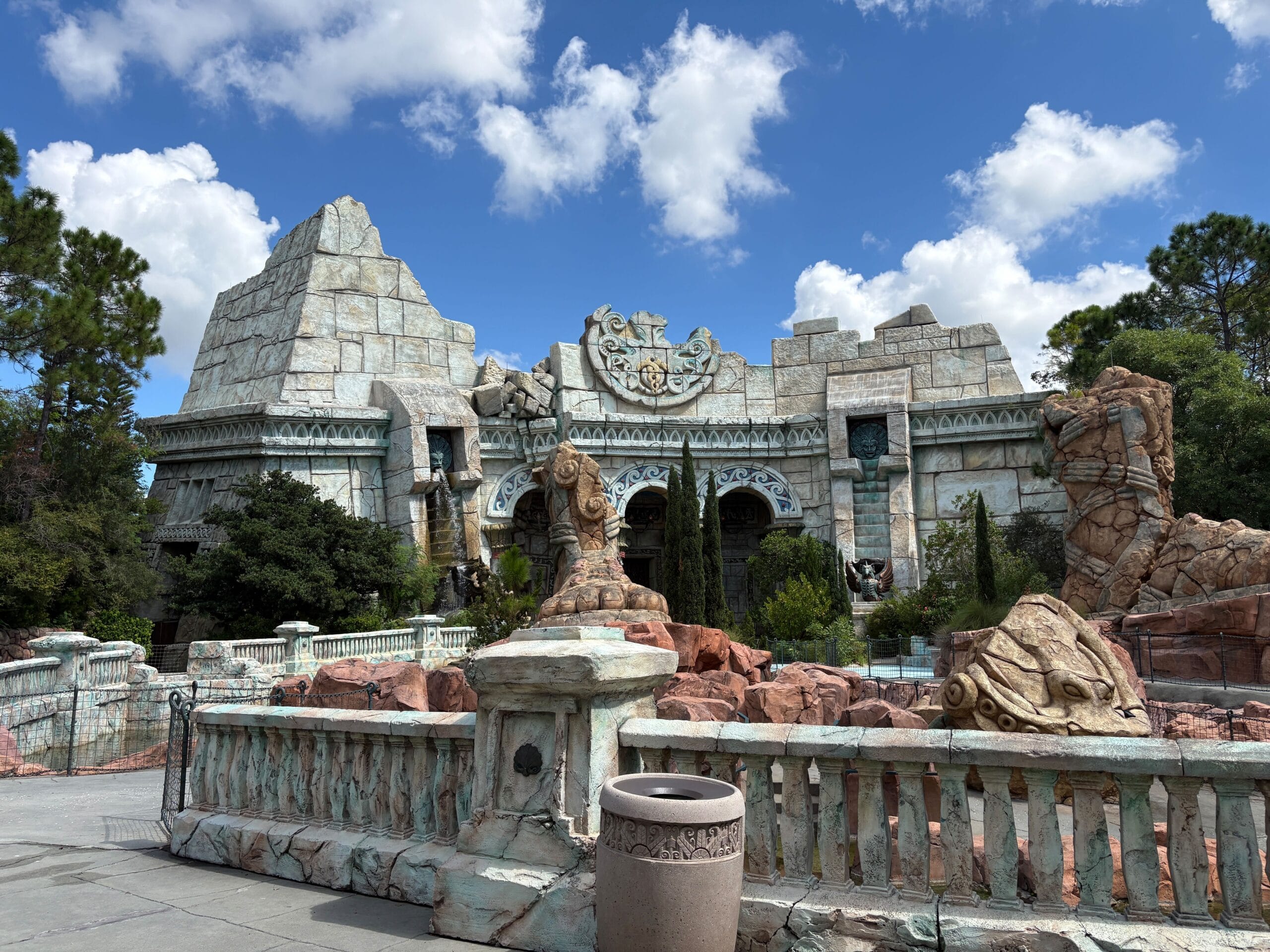 Ancient temple-like building with stone statues, rocky landscape, and blue sky with clouds in the background.