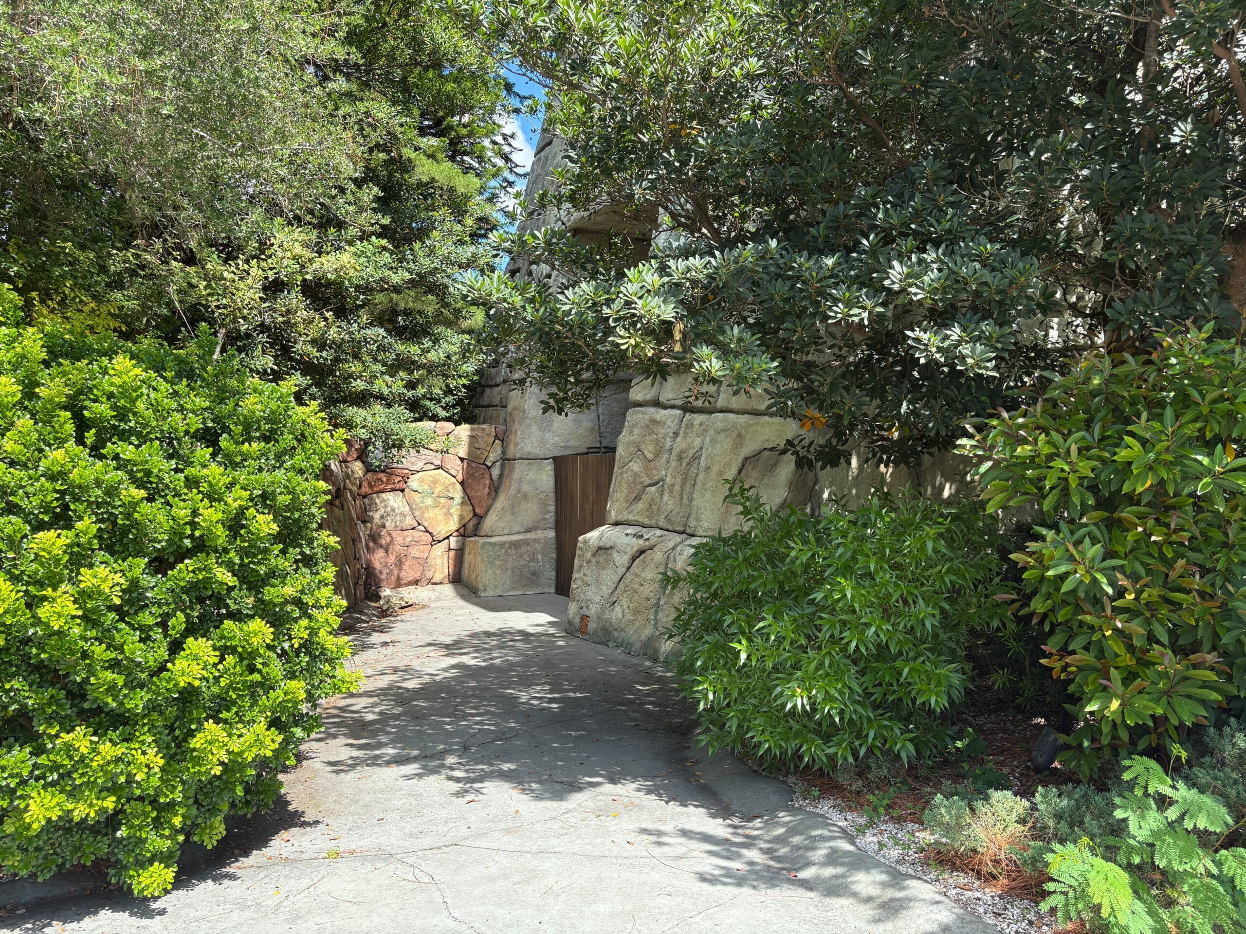 A narrow stone pathway surrounded by lush green bushes and trees, leading to a wooden gate.
