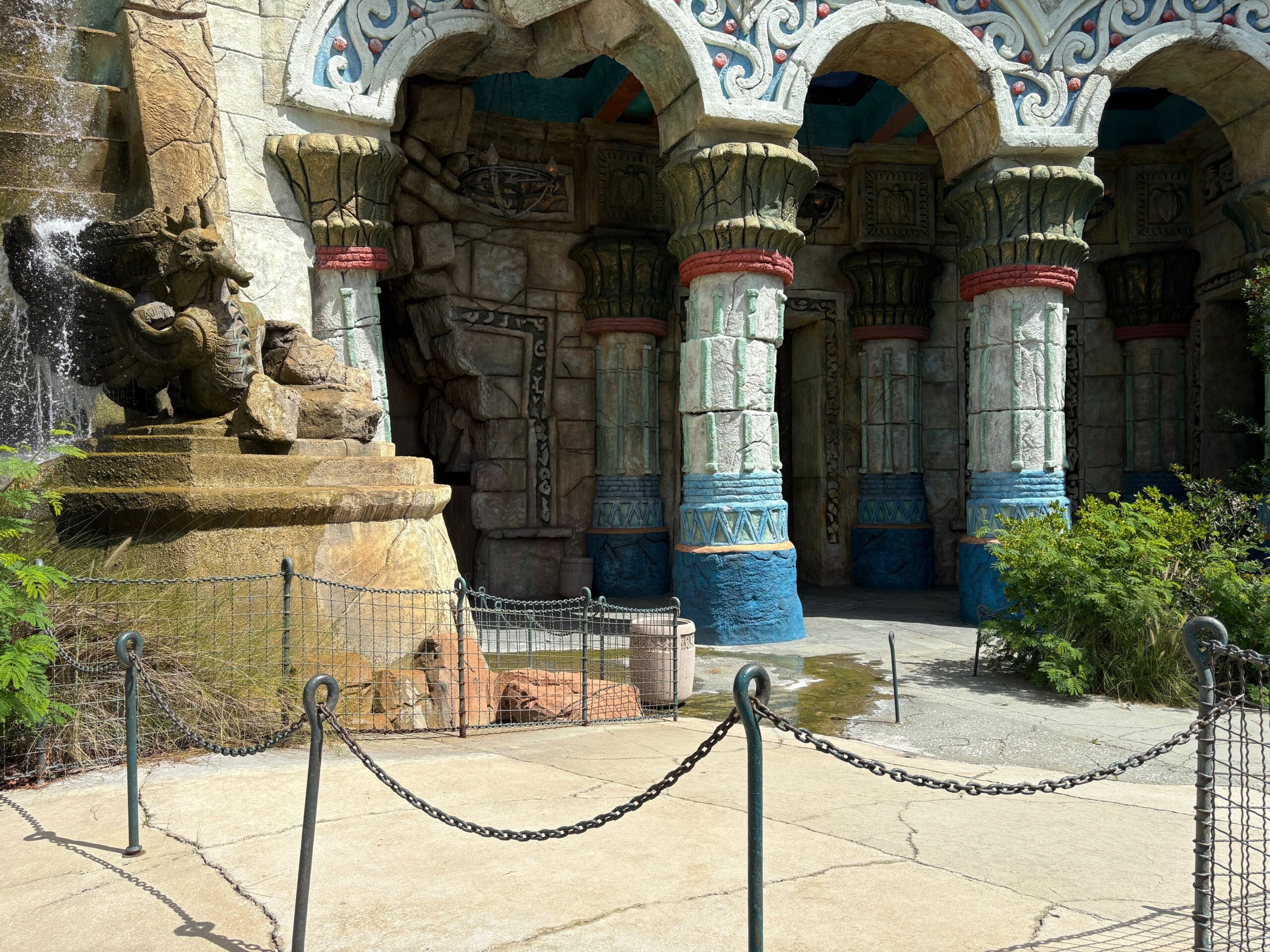 Stone columns with colorful carvings stand beside a dragon statue and waterfall at an ornate building entrance.