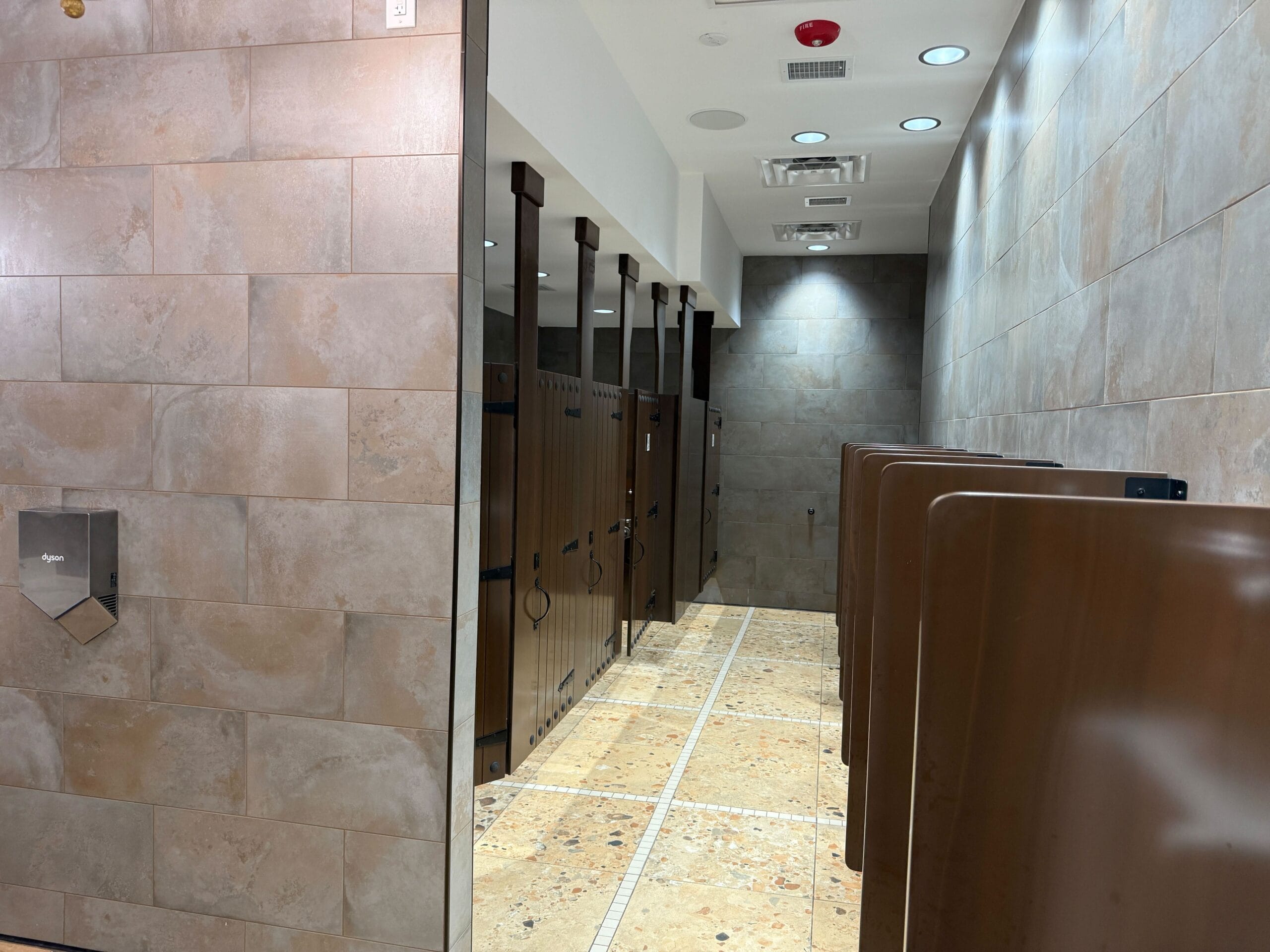 A clean Port of Entry Restrooms with tan tiled walls, brown stall doors, and urinals lined up on the right.