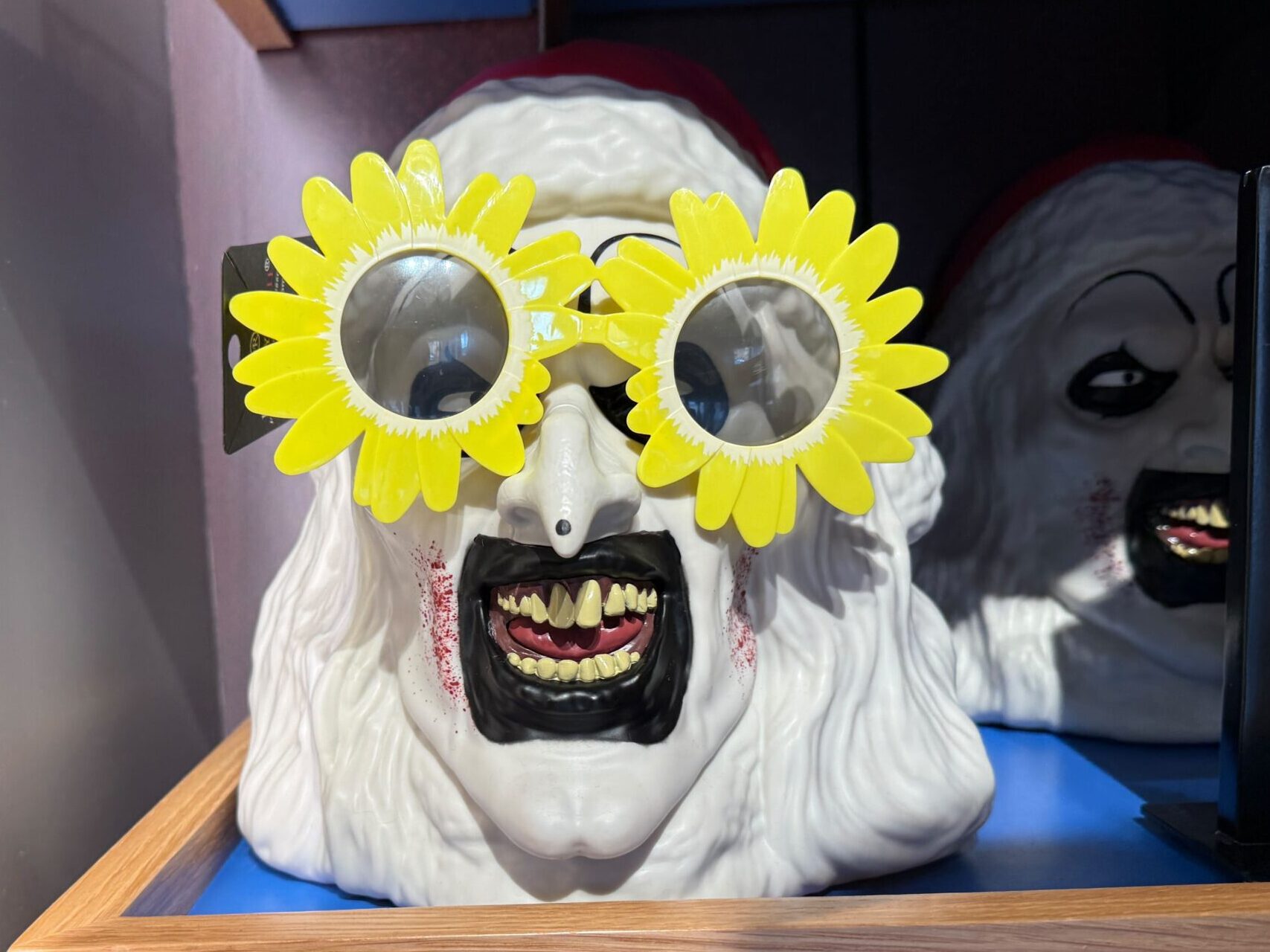 A scary clown mask with Terrifier Sunflower Glasses is displayed on a wooden shelf—perfect for HHN 2025.
