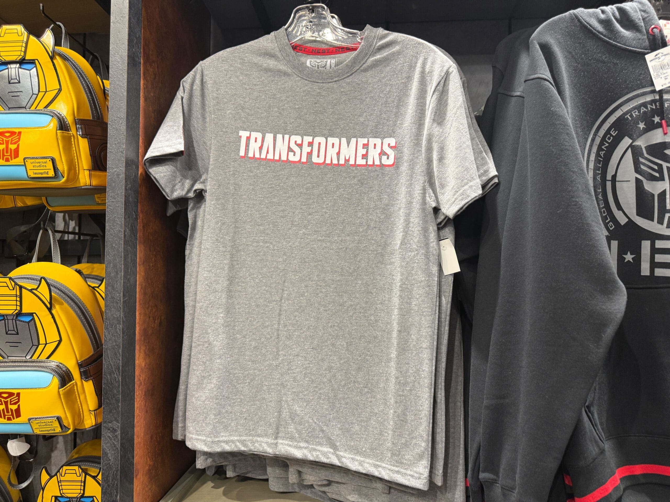 New Transformers NEST Merchandise with "TRANSFORMERS" in white and red letters hanging on a rack next to Transformers-themed bags.