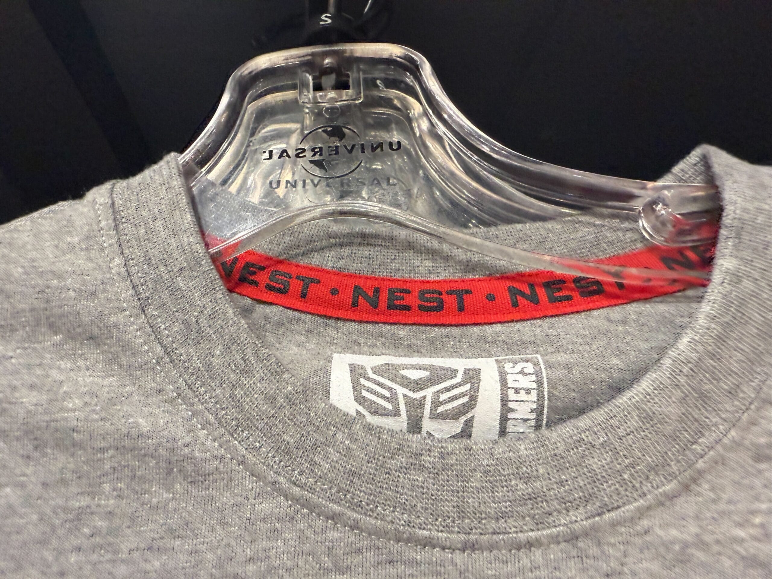 New Transformers NEST Merchandise T-shirt on a hanger with a red collar tag reading "NEST" repeatedly in black letters.