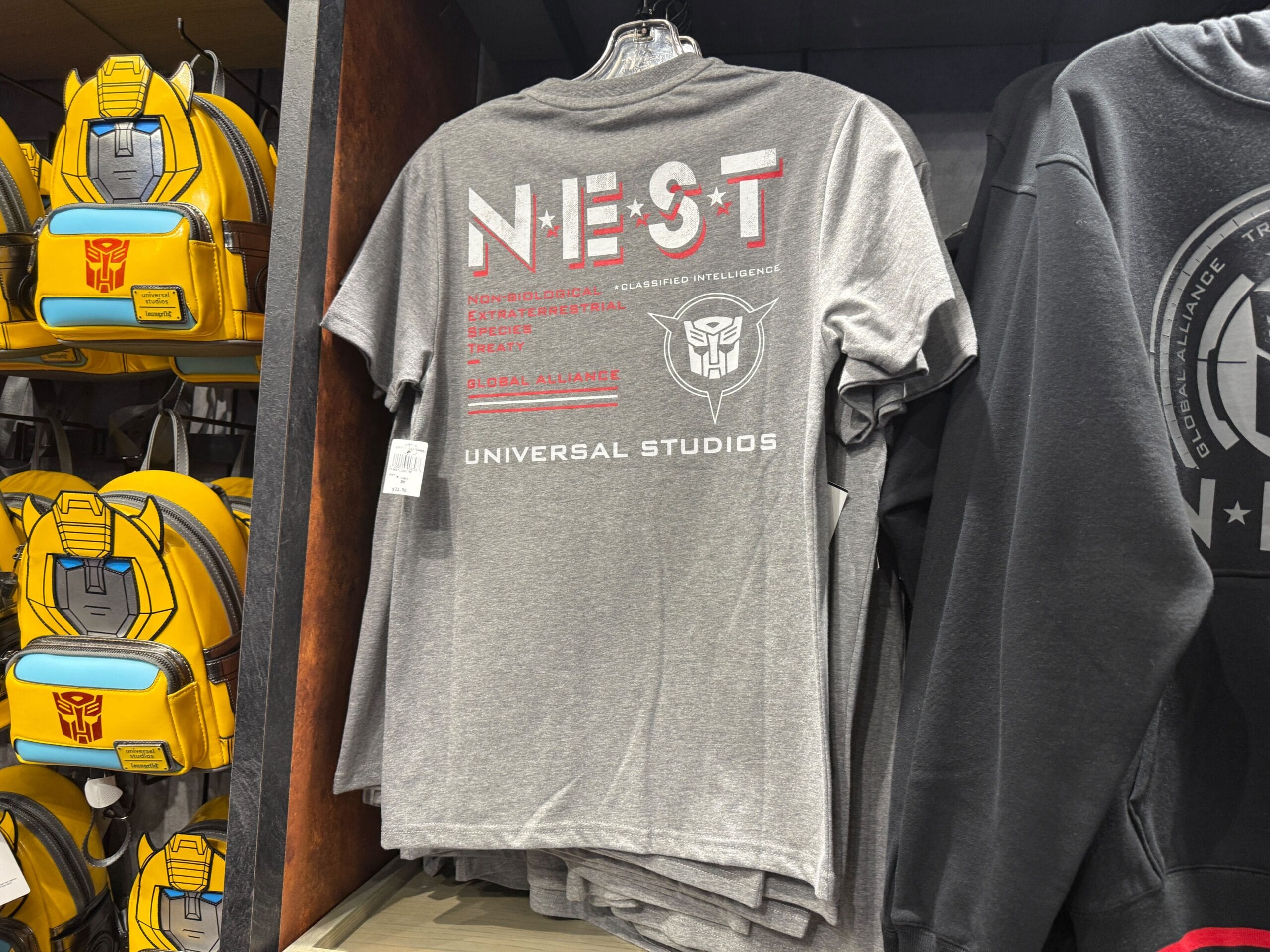 Gray NEST t-shirt with Transformers logo displayed on a shelf at Universal Studios, next to yellow Transformers backpacks.