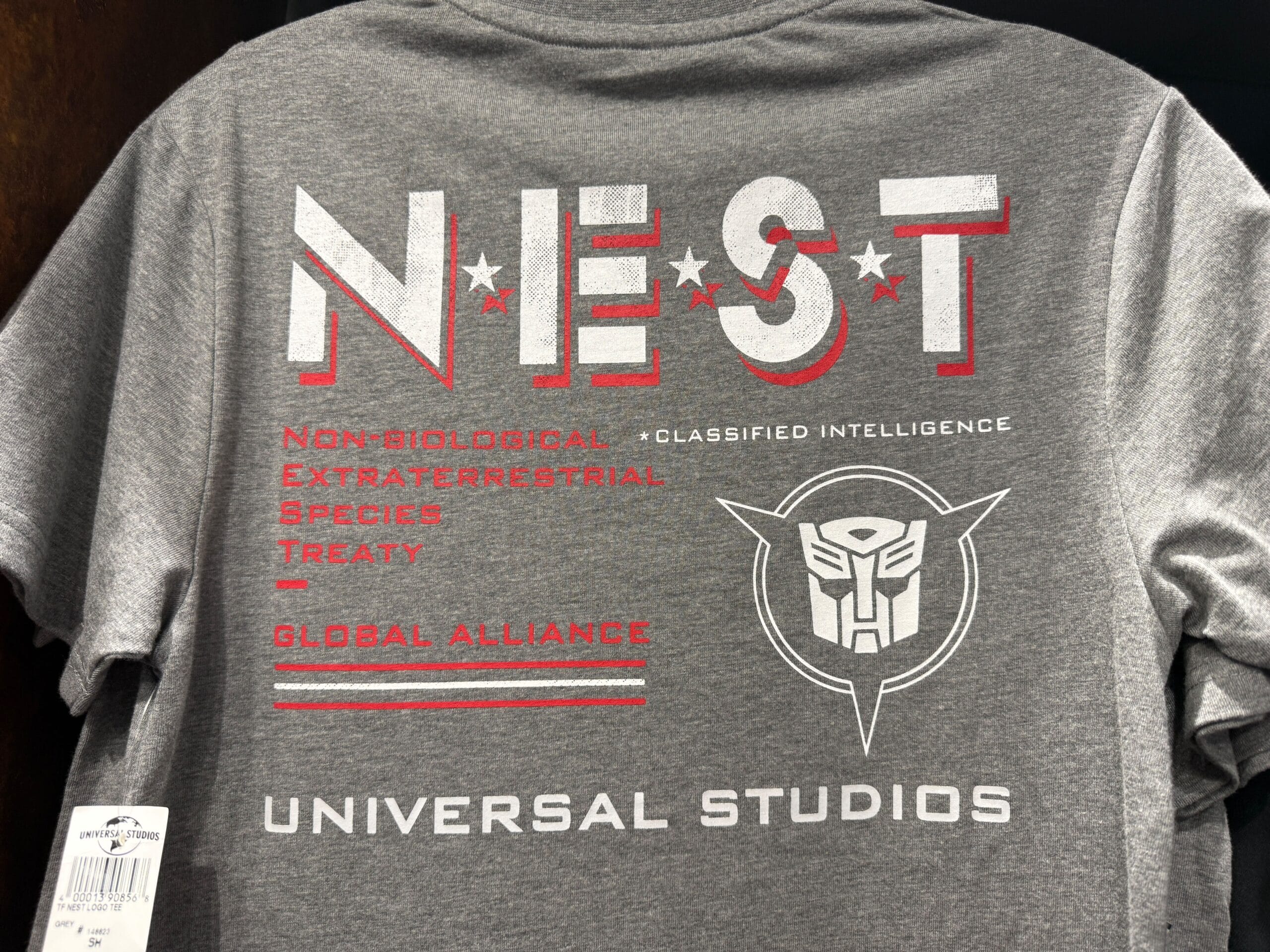 Gray Universal Studios T-shirt with NEST logo, Transformers symbol, and related text in white and red.