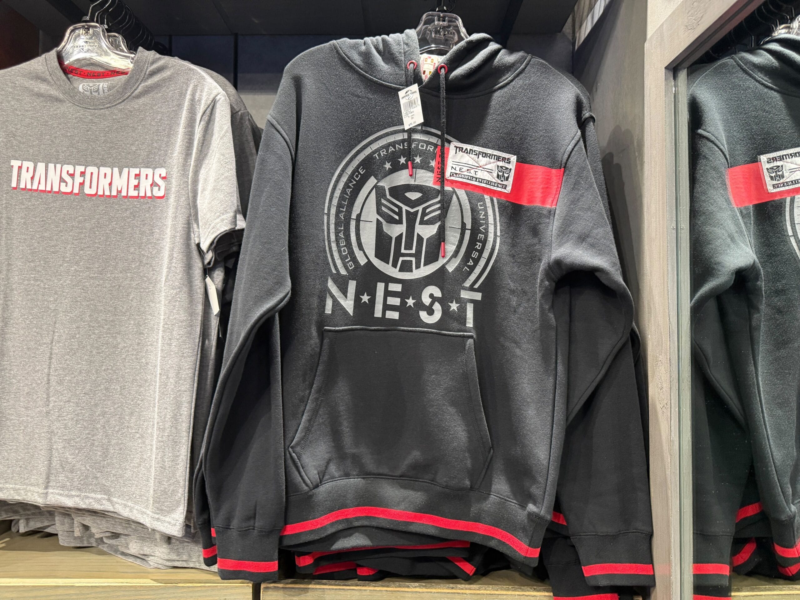 A black Transformers NEST hoodie with red accents displayed next to a gray Transformers t-shirt in a store.